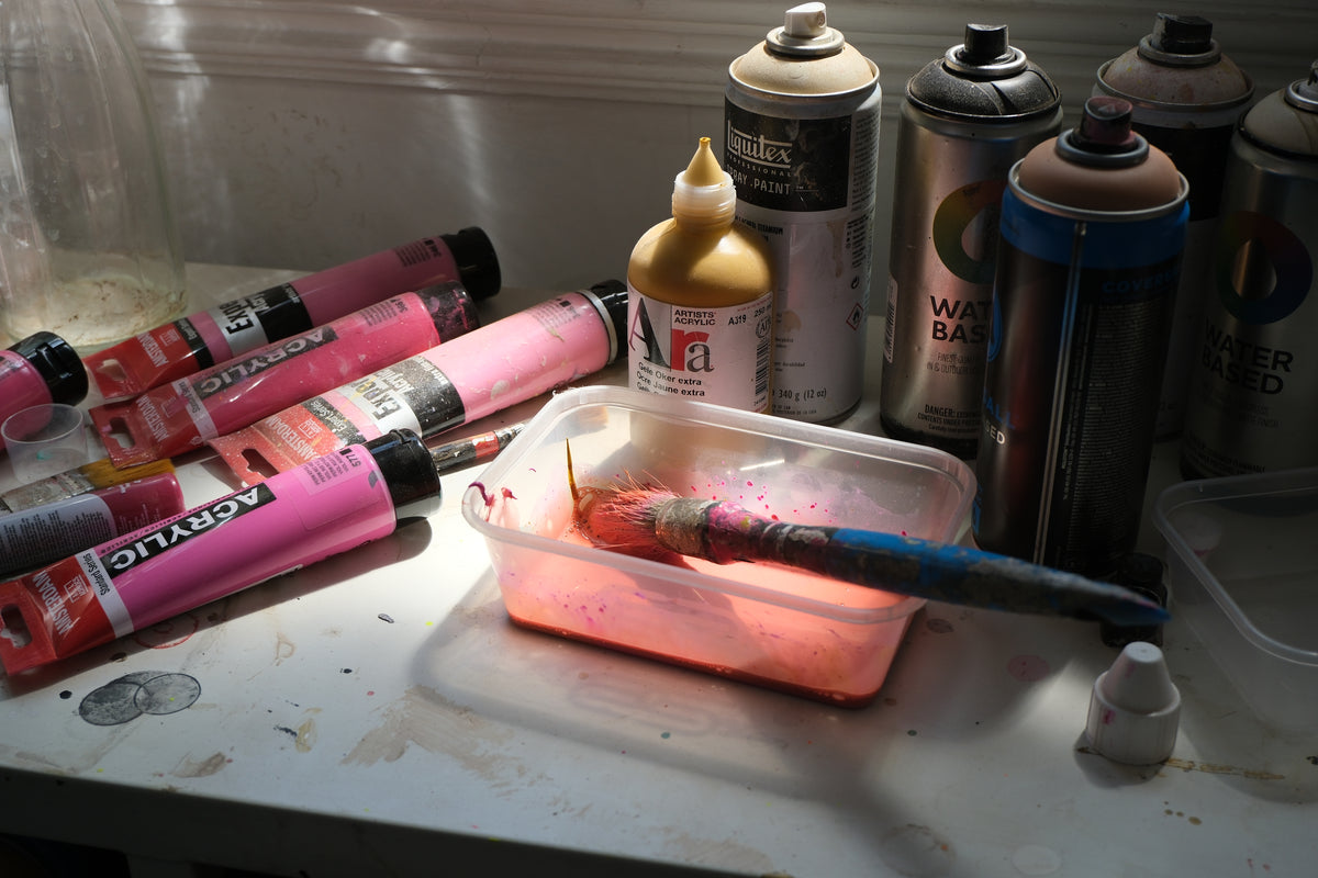 A paintbrush in a stained plastic container, acrylic tubes, and spray paint cans on a messy white surface.