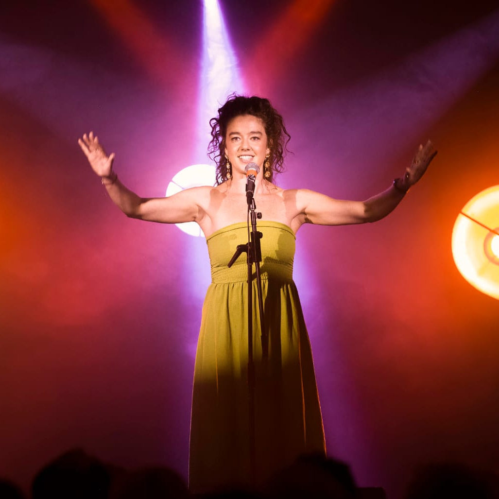 Elle Dillon-Reams stands on a stage with her arms outstretched, wearing a strapless olive green dress and holding a microphone. She is illuminated by dramatic purple and orange stage lights, with two glowing circular lights visible on either side.