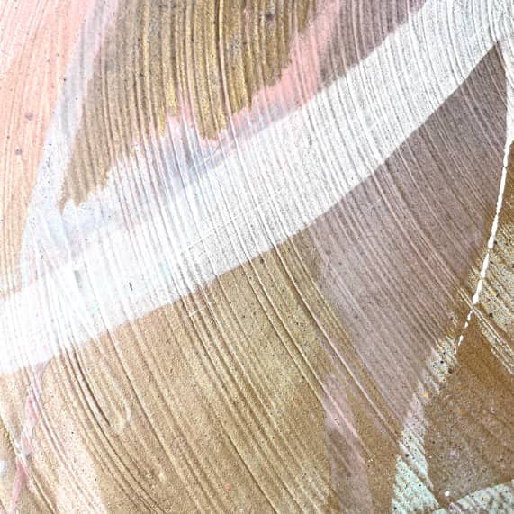 The "Just Be" painting by Kate Mayer showcases overlapping brushstrokes in shades of brown, white, and pink, utilizing acrylic and spray paint to achieve a textured, layered effect.
