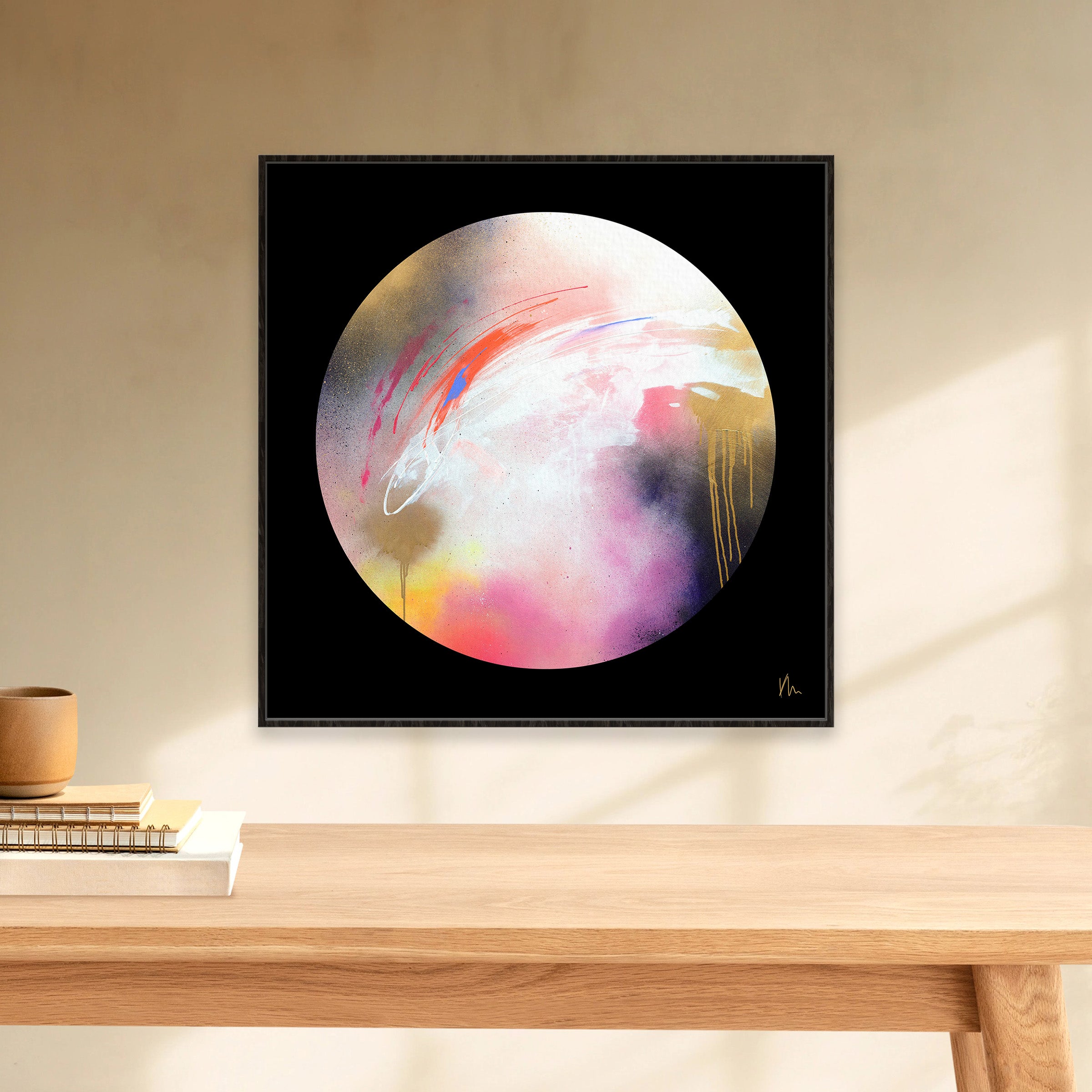 The Dream Starts Here," a framed abstract circular artwork by Kate Mayer, features vibrant colors and hangs on a beige wall above a wooden shelf. The shelf holds a stack of books and a cup. The vibrant hues and modern design of this piece create a captivating focal point in any room.