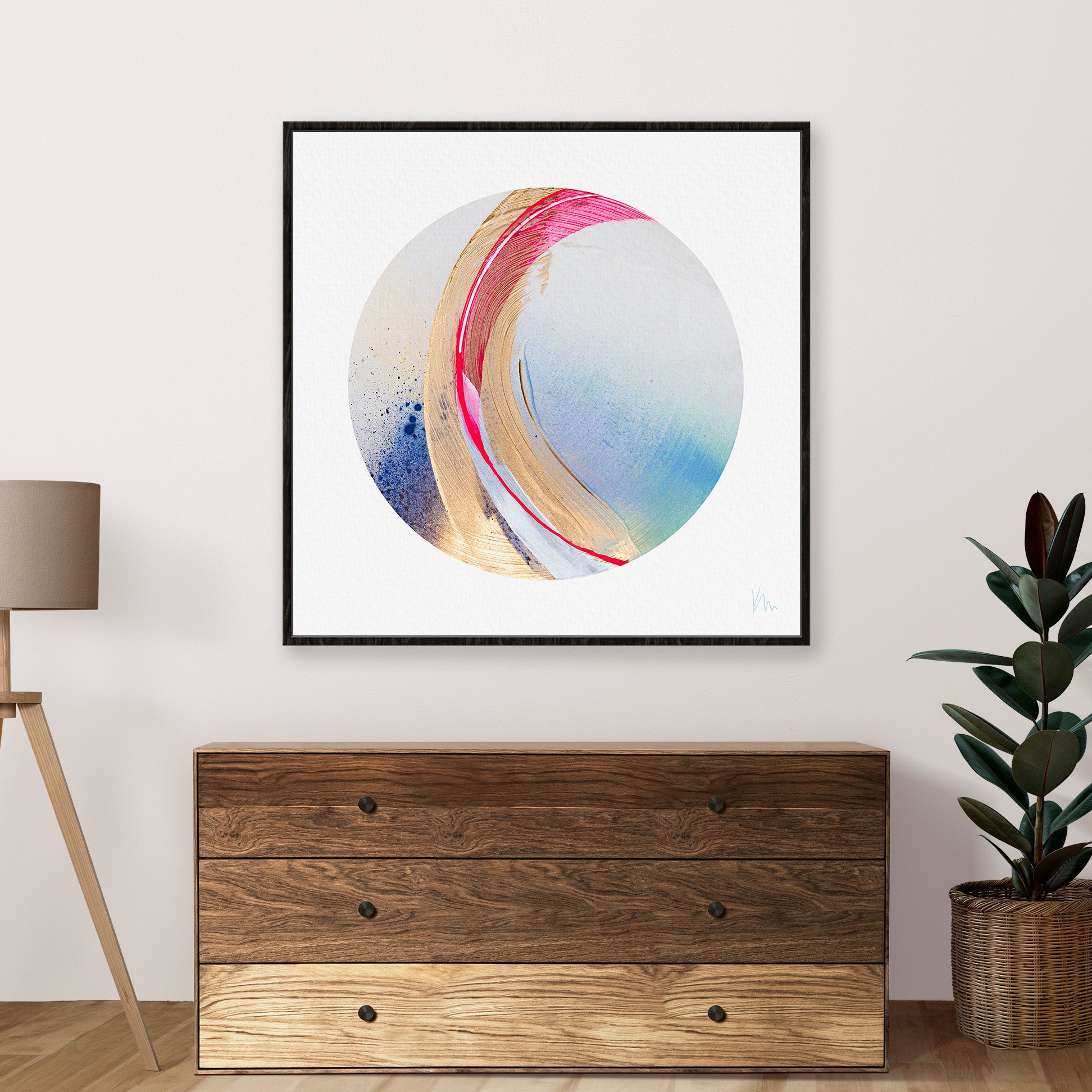 The "Be Radiant" framed original painting by Kate Mayer, featuring a dynamic arrangement of multicolored circular designs, is placed above a wooden dresser accompanied by a lamp and plant.