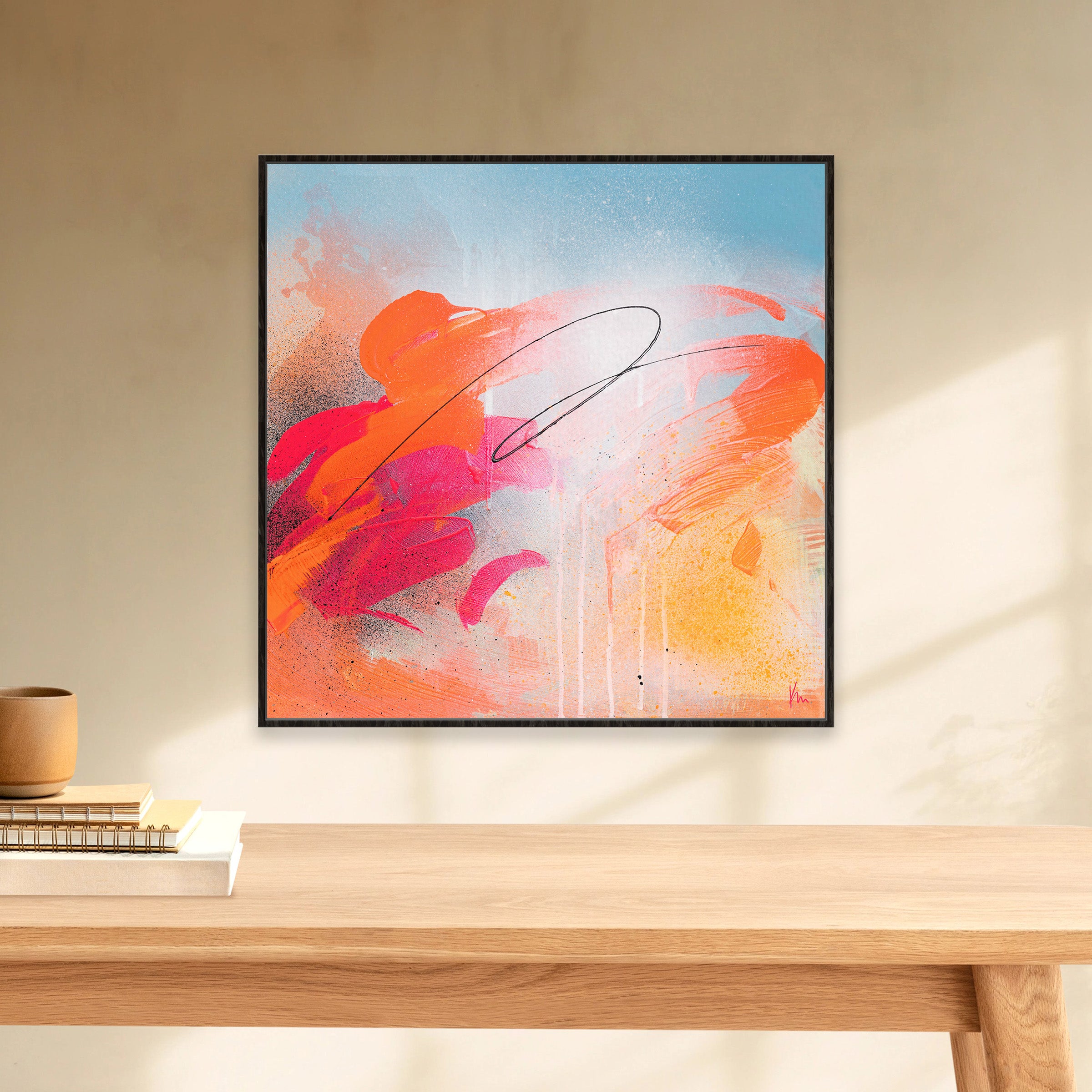 The "Dance With Me" framed abstract painting by Kate Mayer, featuring vibrant orange, red, and blue brushstrokes, adorns the wall above a wooden table with books and a small pot.