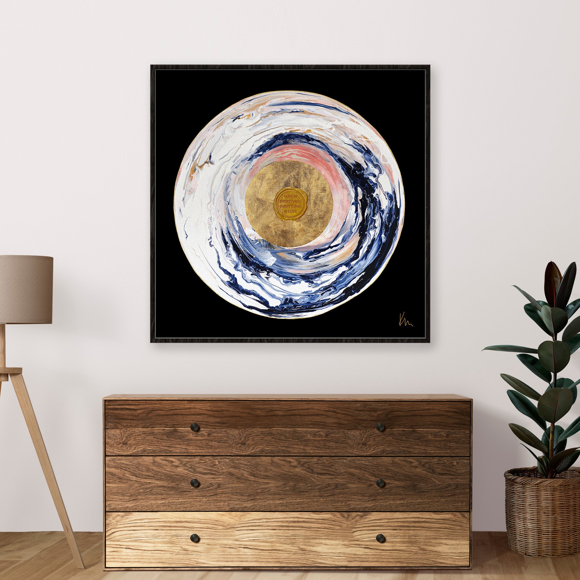 The abstract circular painting, "Love Sublime" by Kate Mayer, features a captivating gold center and swirling colors on a black backdrop. It elegantly graces the space above a wooden dresser adorned with lush greenery.