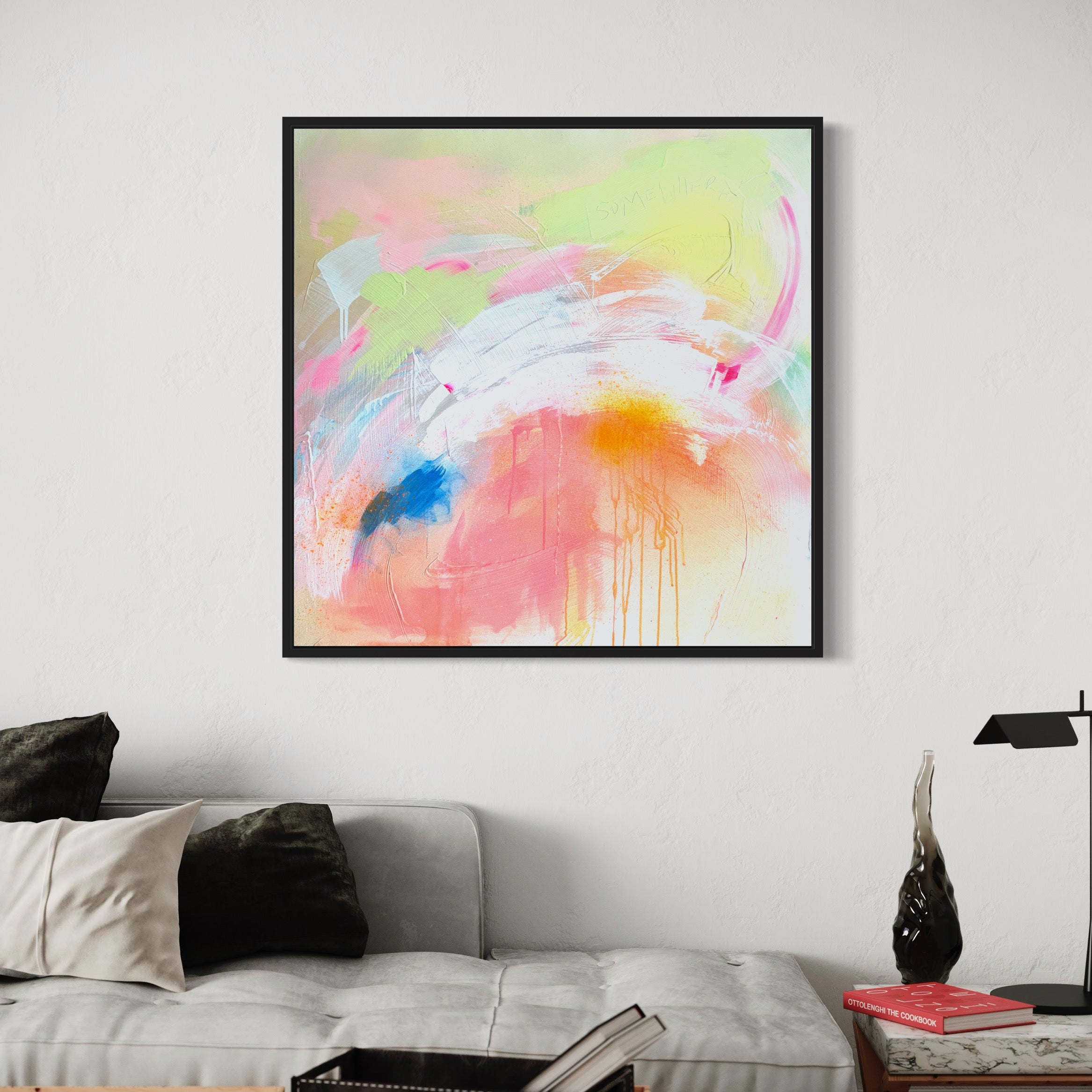 A framed original painting titled "Pure Joy" by Kate Mayer, showcasing abstract designs in pastel colors and created using acrylic and spray paint on canvas, is displayed above a modern sofa adorned with black and white cushions. The ensemble is completed with a small side table holding books.