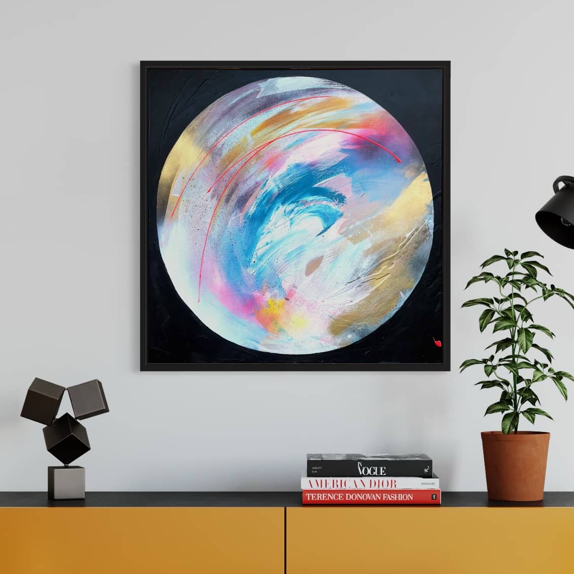 The framed "Speak Your Truth - Original Painting" by Kate Mayer features a colorful circular design and is displayed on a wall above a yellow cabinet adorned with books, a plant, and geometric decor.
