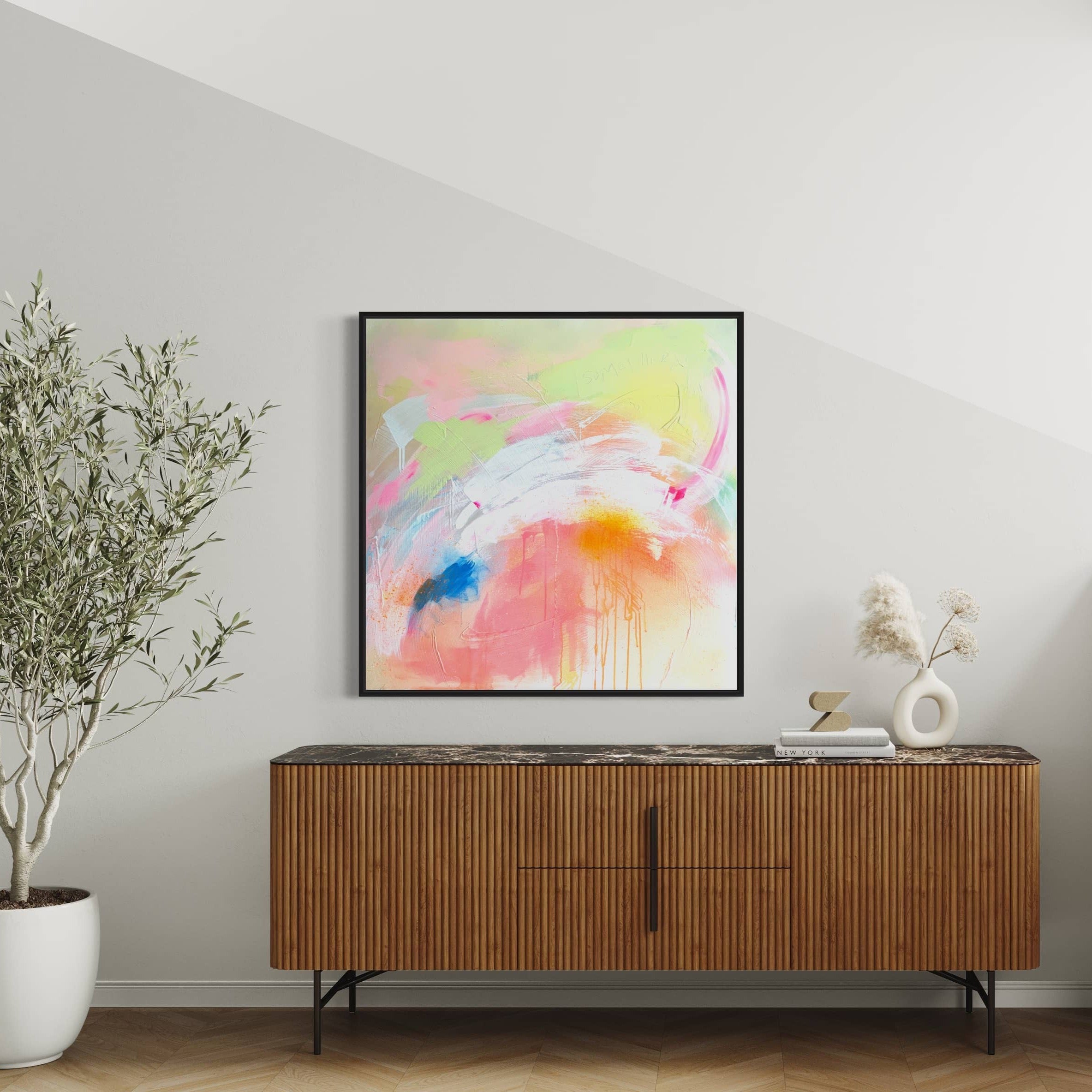 Pure Joy - Original Painting by Kate Mayer, framed in a black wooden float frame, hangs above a wooden sideboard. The vibrant colors of the acrylic and spray paint on canvas complement the potted plant and decorative items placed below.