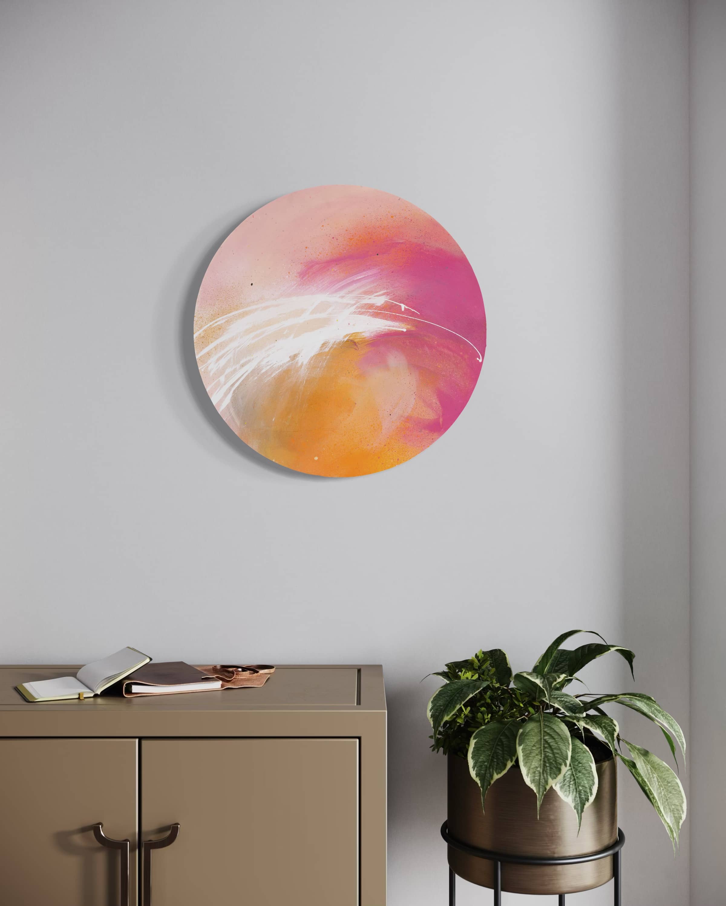 Shine On by Kate Mayer, a round abstract pink and orange artwork on plywood, displayed above a cabinet with a plant nearby.