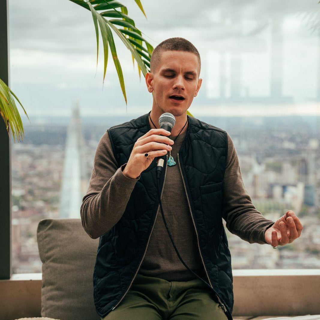 Tor Njamo sits on a couch in a high-rise apartment, holding a microphone and singing. He is wearing a black quilted vest over a brown long-sleeve shirt and olive green pants.
