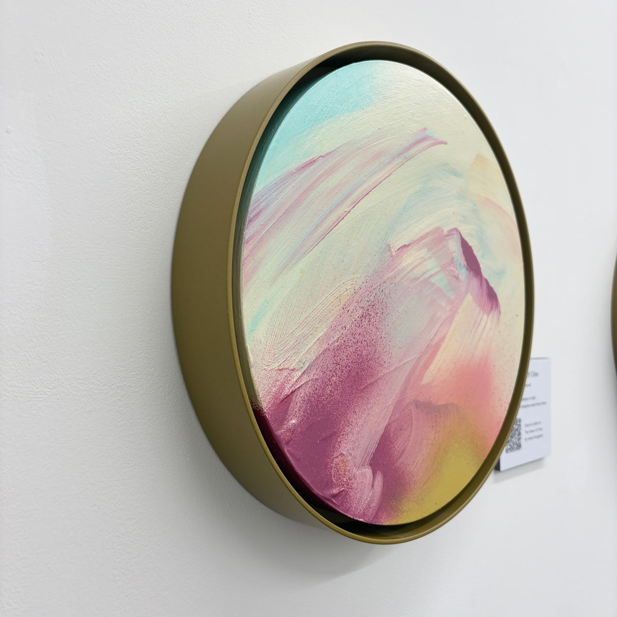 The Power Of One by Kate Mayer: round acrylic abstract in pastel hues, gold metal frame, on white wall.