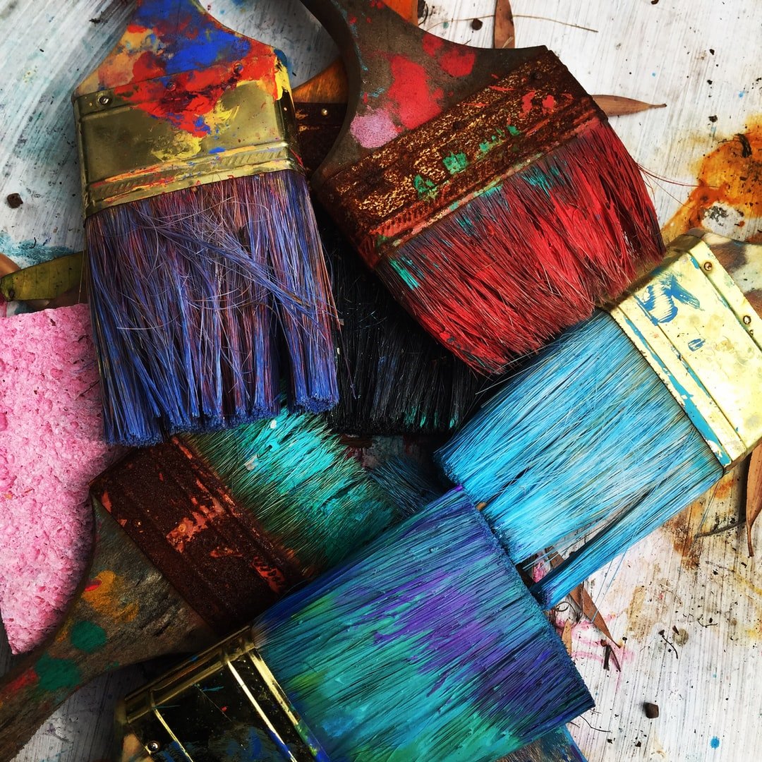 Five paintbrushes with vibrant, well-loved bristles lie overlapping on a textured surface, showcasing the artistic remnants of Kate Mayer's Self Expression Workshop, with splatters of pink, blue, and red paint.