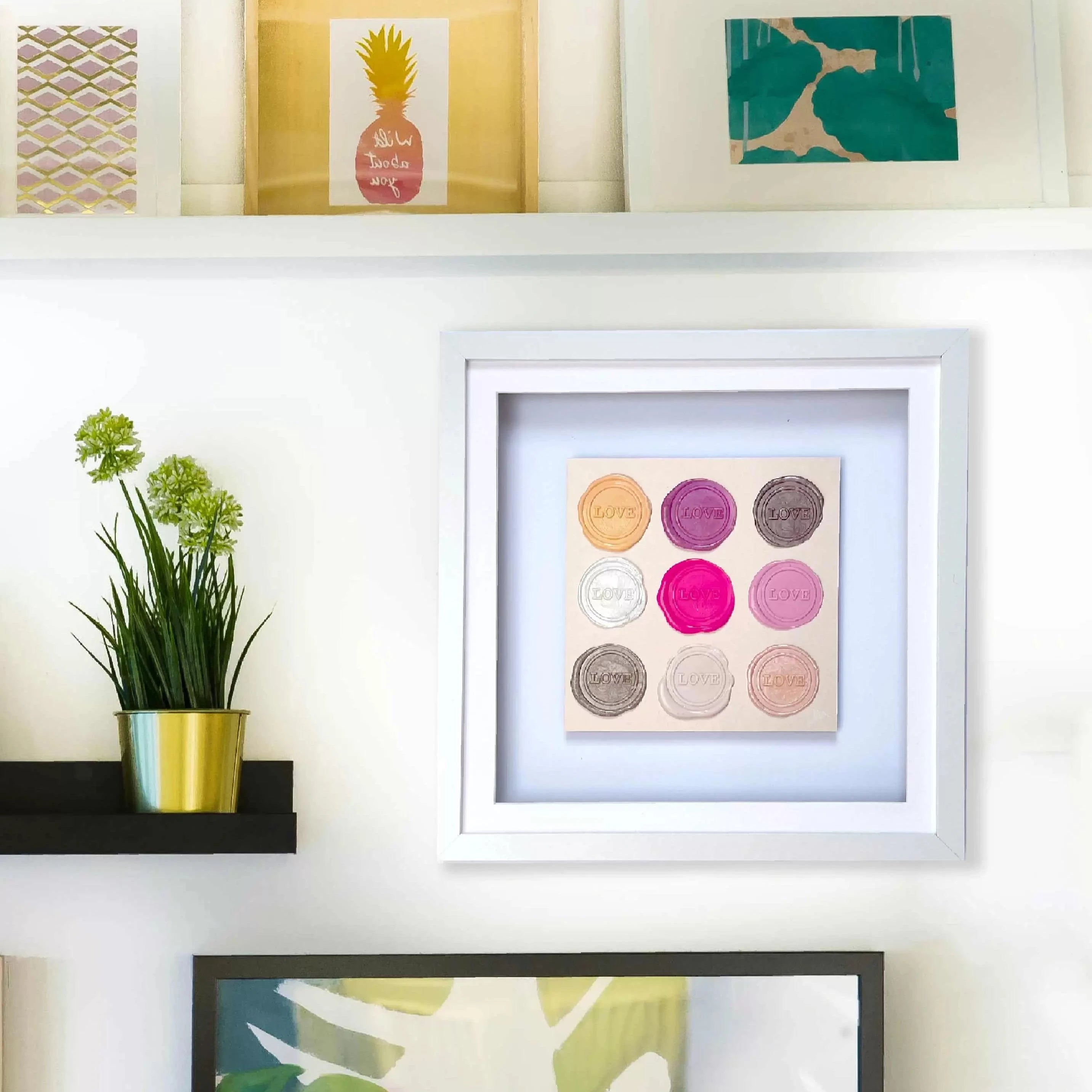 White shelves with framed original art and a potted plant. The central piece, "Sealed Intention - Love is Love" by Kate Mayer, signed by the artist, showcases a grid of colored circles.