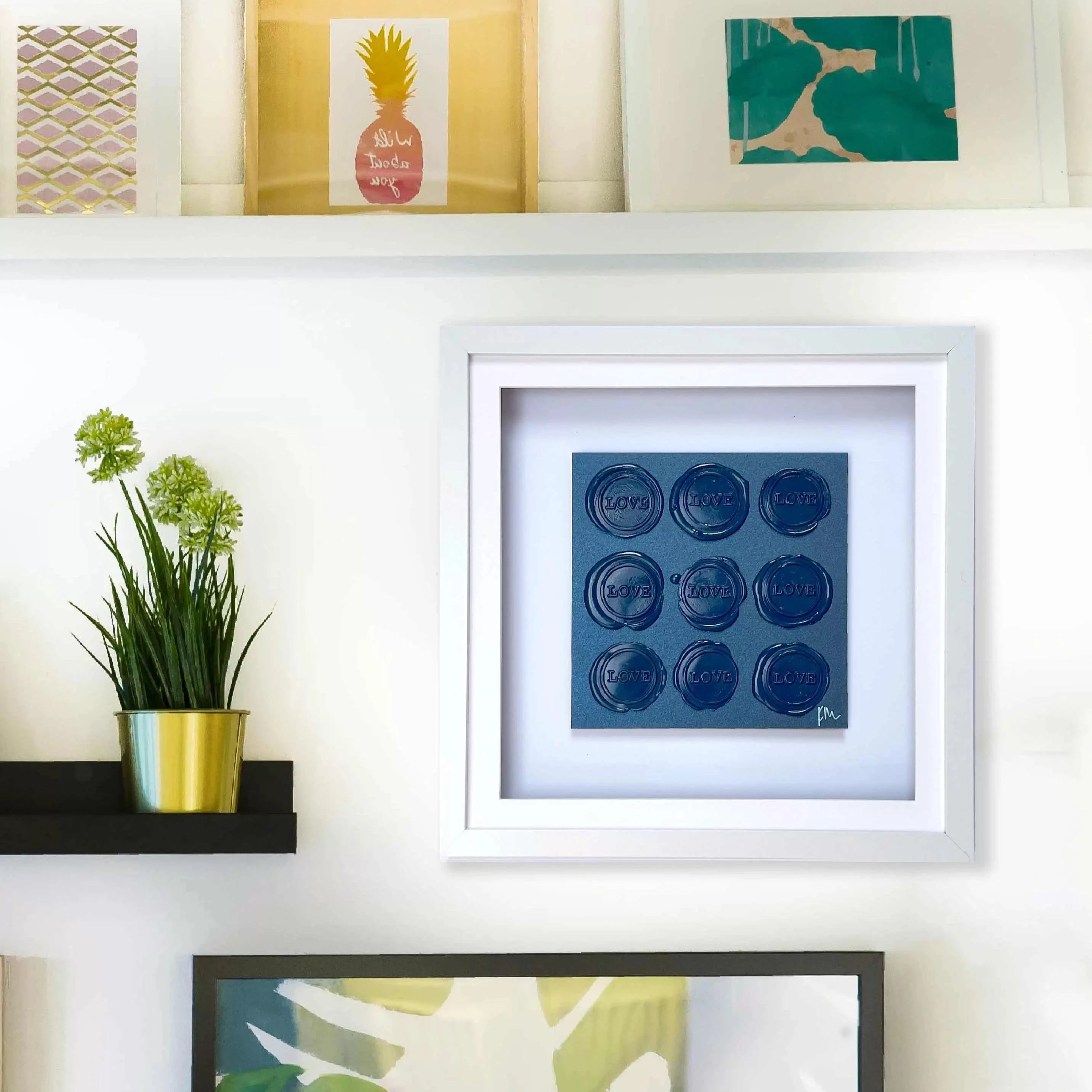 The white shelf is beautifully adorned with framed artwork and a potted plant, complemented by vibrant prints and abstract designs. Explore the uniqueness of "Sealed Intention - Love is Love" by Kate Mayer, featuring original art signed by the artist to add a personal touch to your space.