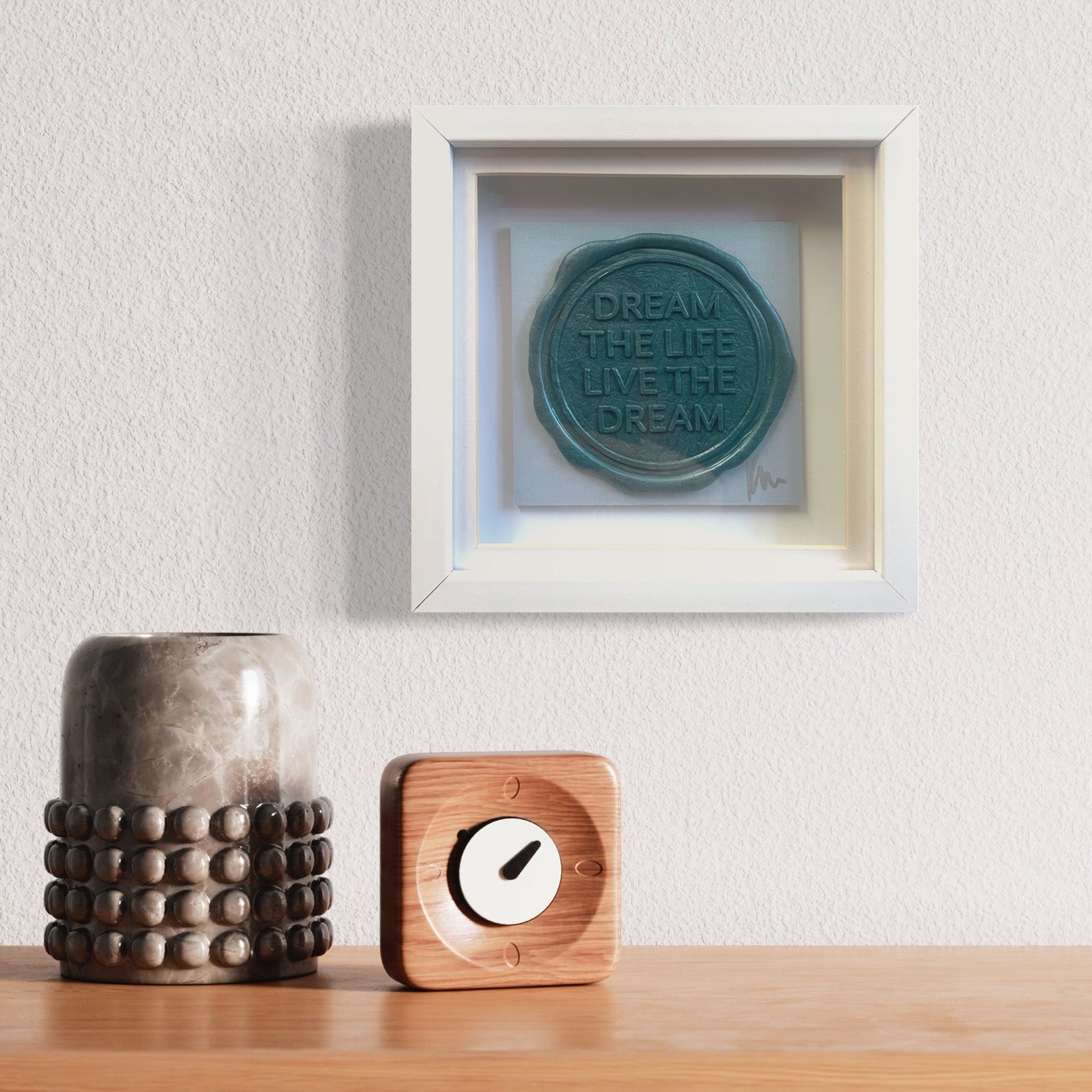 A framed quote from Kate Mayer's "Sealed Intention to Live The Dream" hangs above a wooden shelf with a vase, a small wooden clock, and an original art piece nearby.