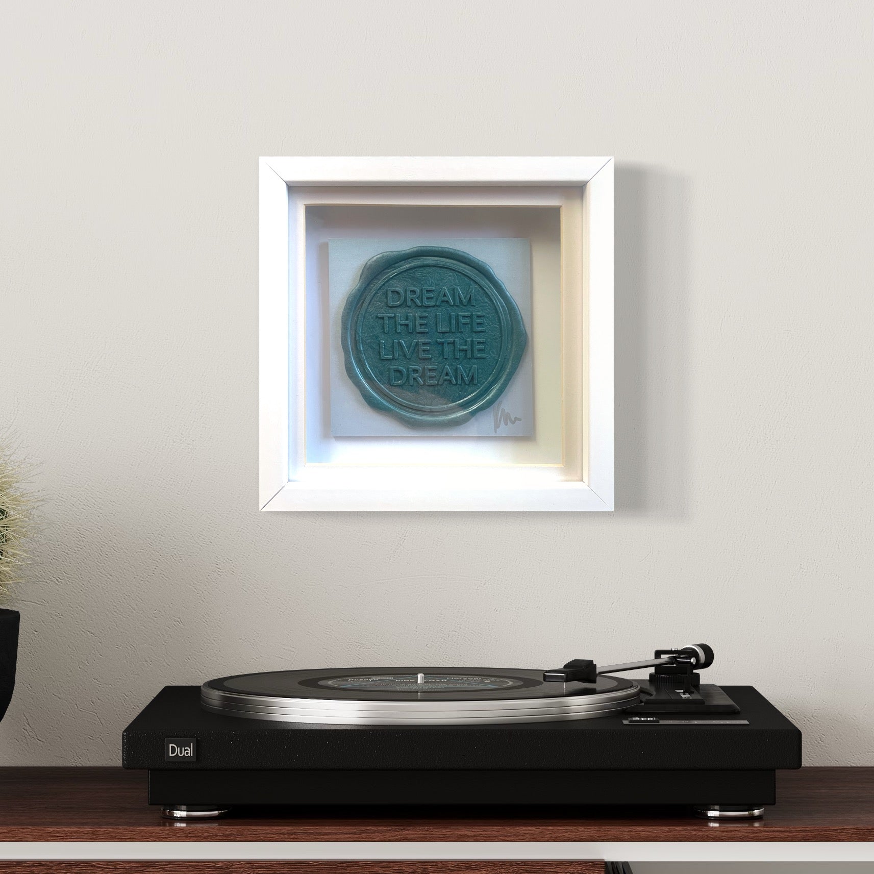 A wall adorned with Kate Mayer's Sealed Intention to Live The Dream artwork in a white shadow frame, accompanied by a vinyl record player on a wooden table below.