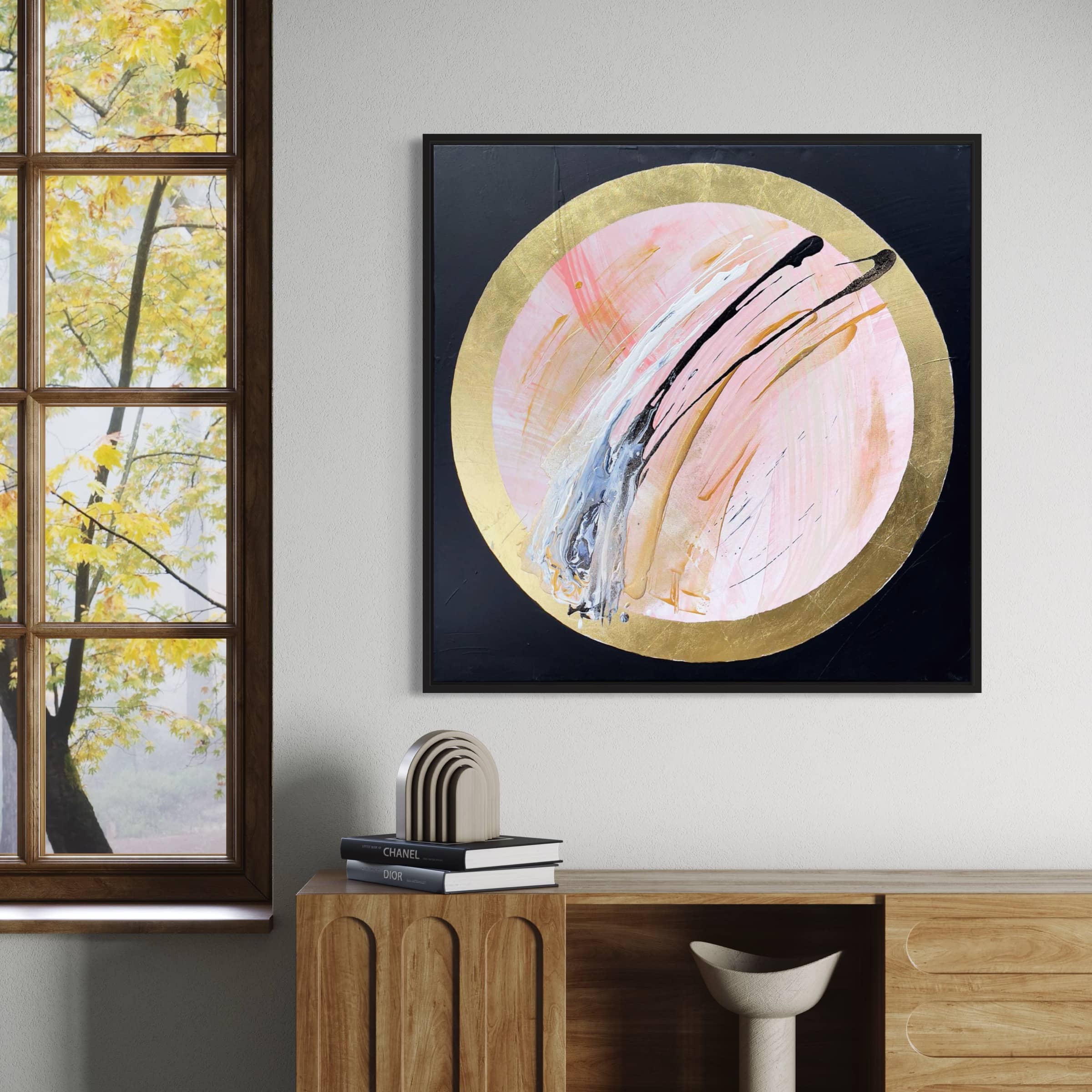 The painting "Let's Go Beyond" by Kate Mayer, featuring a gold-rimmed circle crafted with acrylic and gold leaf, is displayed on a wall beside a window showcasing fall foliage. Below, a cabinet filled with books and decor.