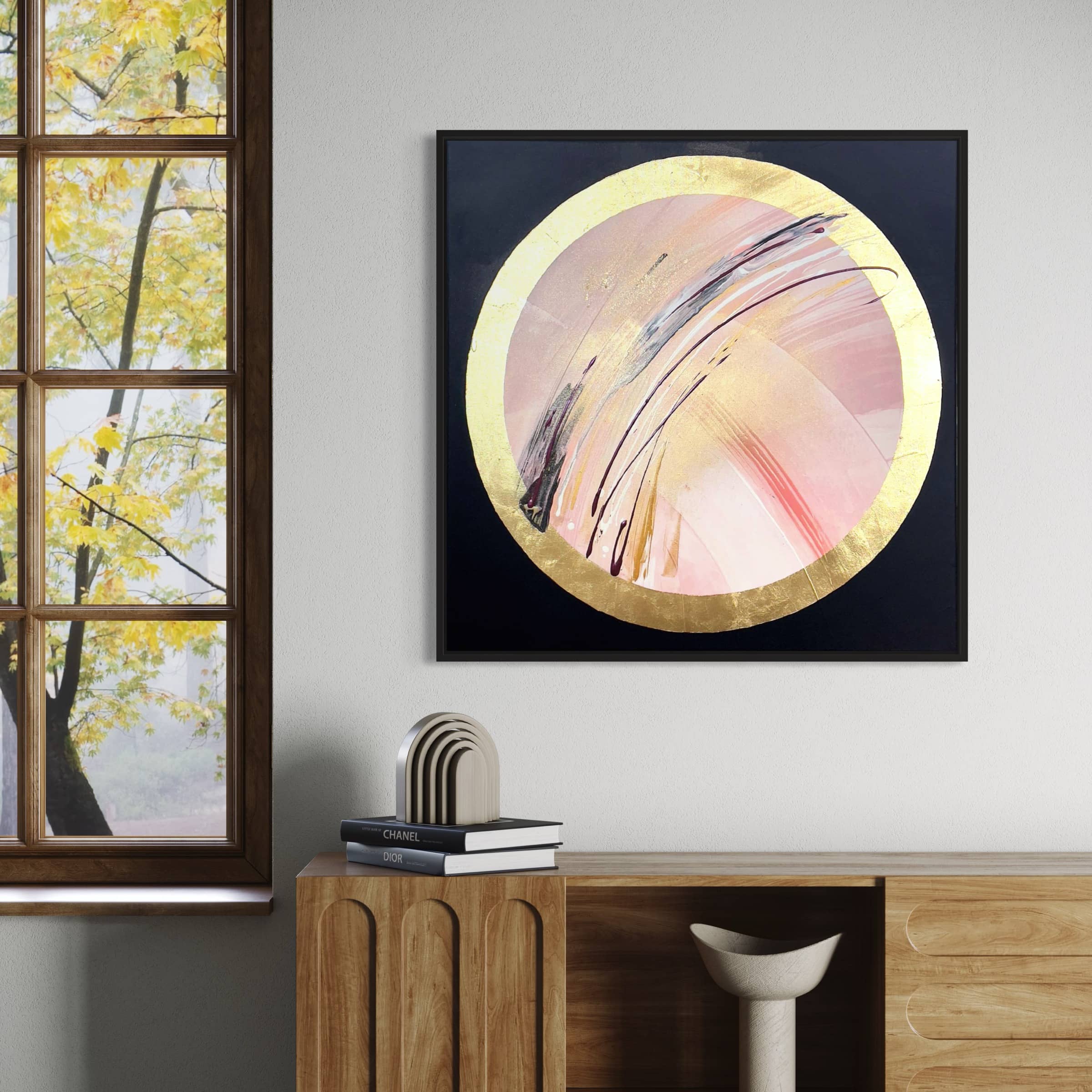 A wooden sideboard with books and decor beneath the framed "Fly With Me" abstract painting by Kate Mayer on the wall, next to a large window showcasing a view of trees.