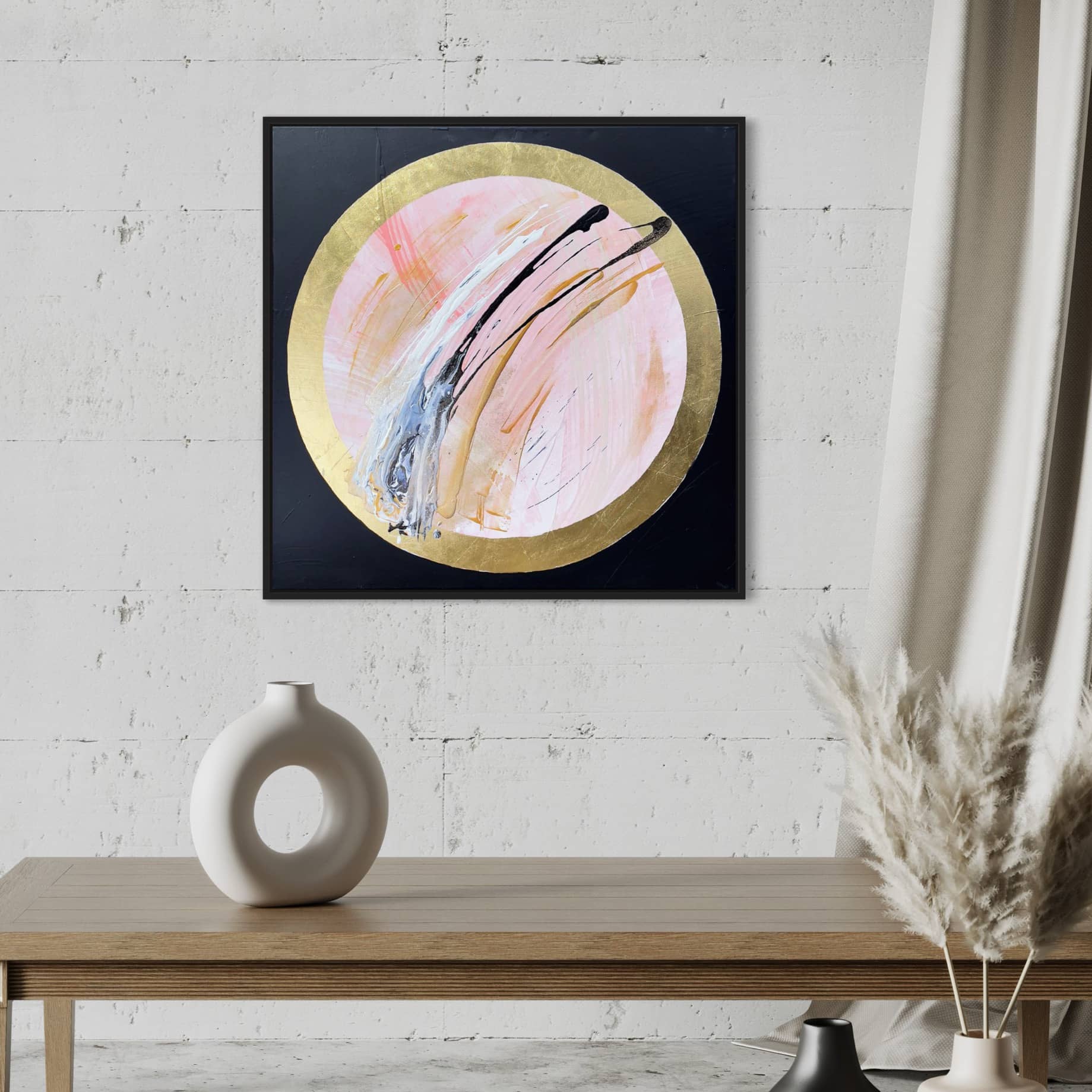 The abstract painting "Let's Go Beyond" by Kate Mayer features a circular design and is displayed on a white wall above a wooden table, which is decorated with a white vase containing a bouquet of pampas grass. The artwork includes acrylic and gold leaf elements, adding a touch of elegance to the minimalist setting.