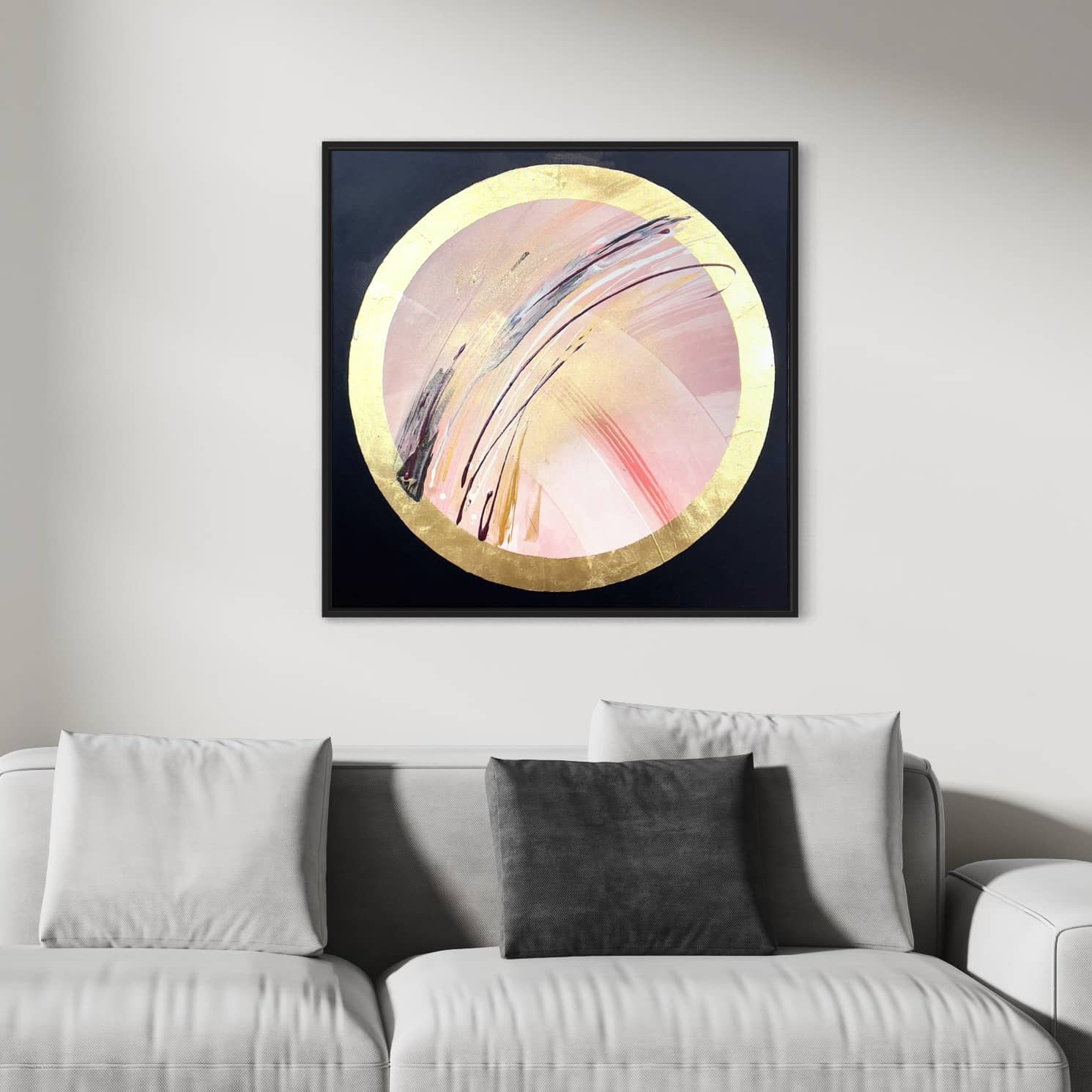 A stunning piece of art titled "Fly With Me" by Kate Mayer, featuring pink hues and gold leaf accents, graces the wall above a light grey couch adorned with one dark grey pillow and two light grey pillows.