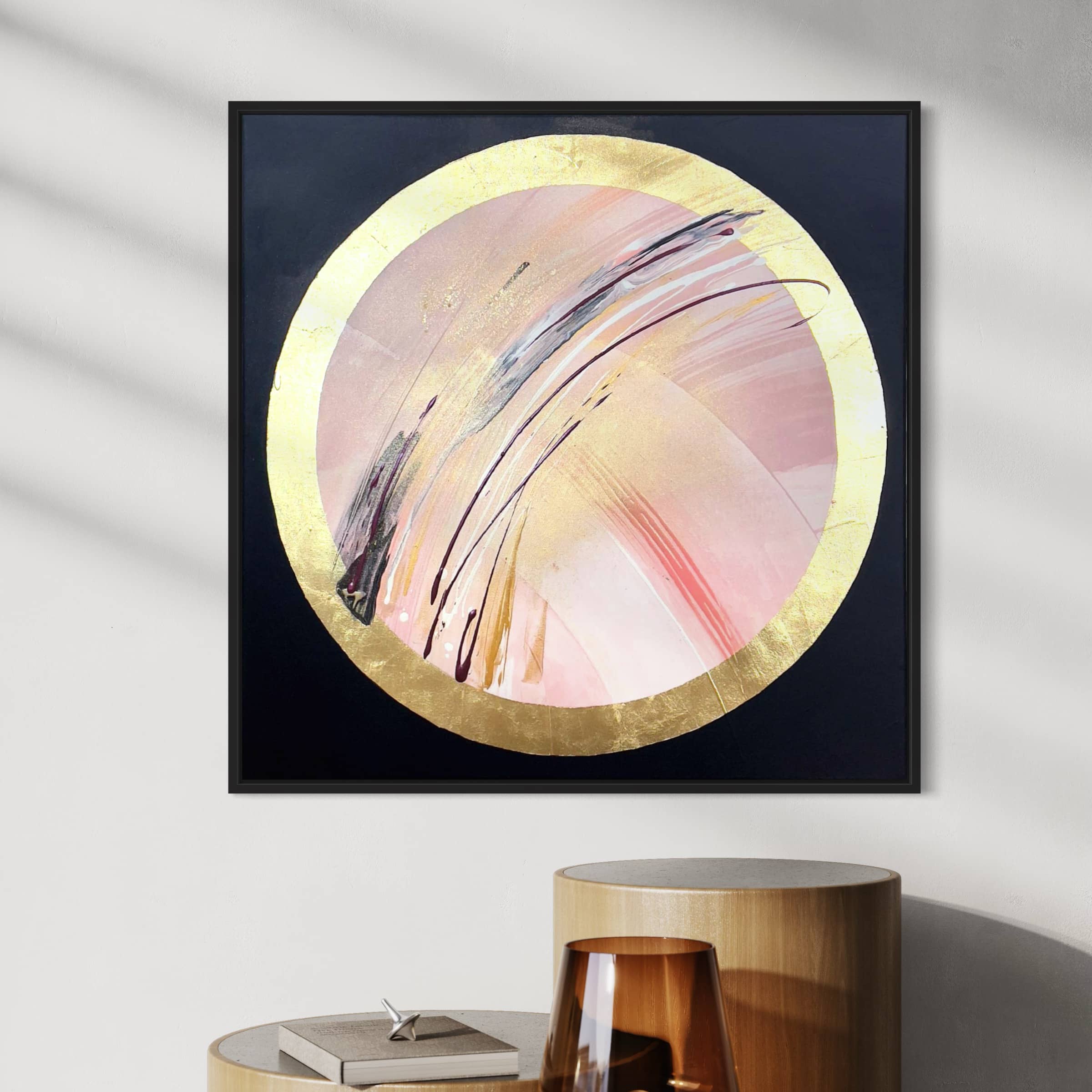 Fly With Me, an acrylic and gold leaf abstract painting by Kate Mayer, features a circular design with gold and pink hues and is mounted on a wall above stacked cylindrical tables.