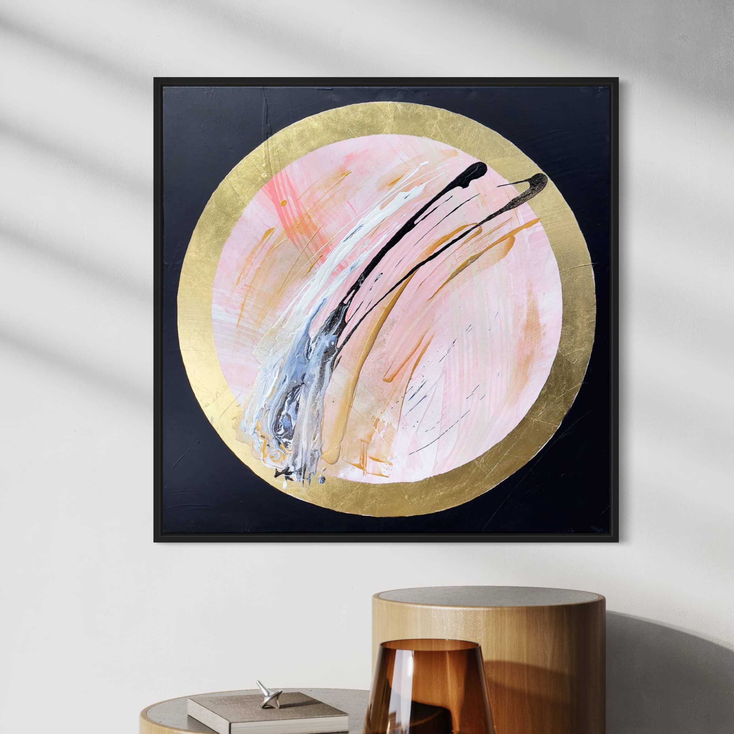 The "Let's Go Beyond" abstract painting by Kate Mayer, featuring a central pink and gold leaf circle with black and light blue brushstrokes, hangs on a wall above a wooden table.