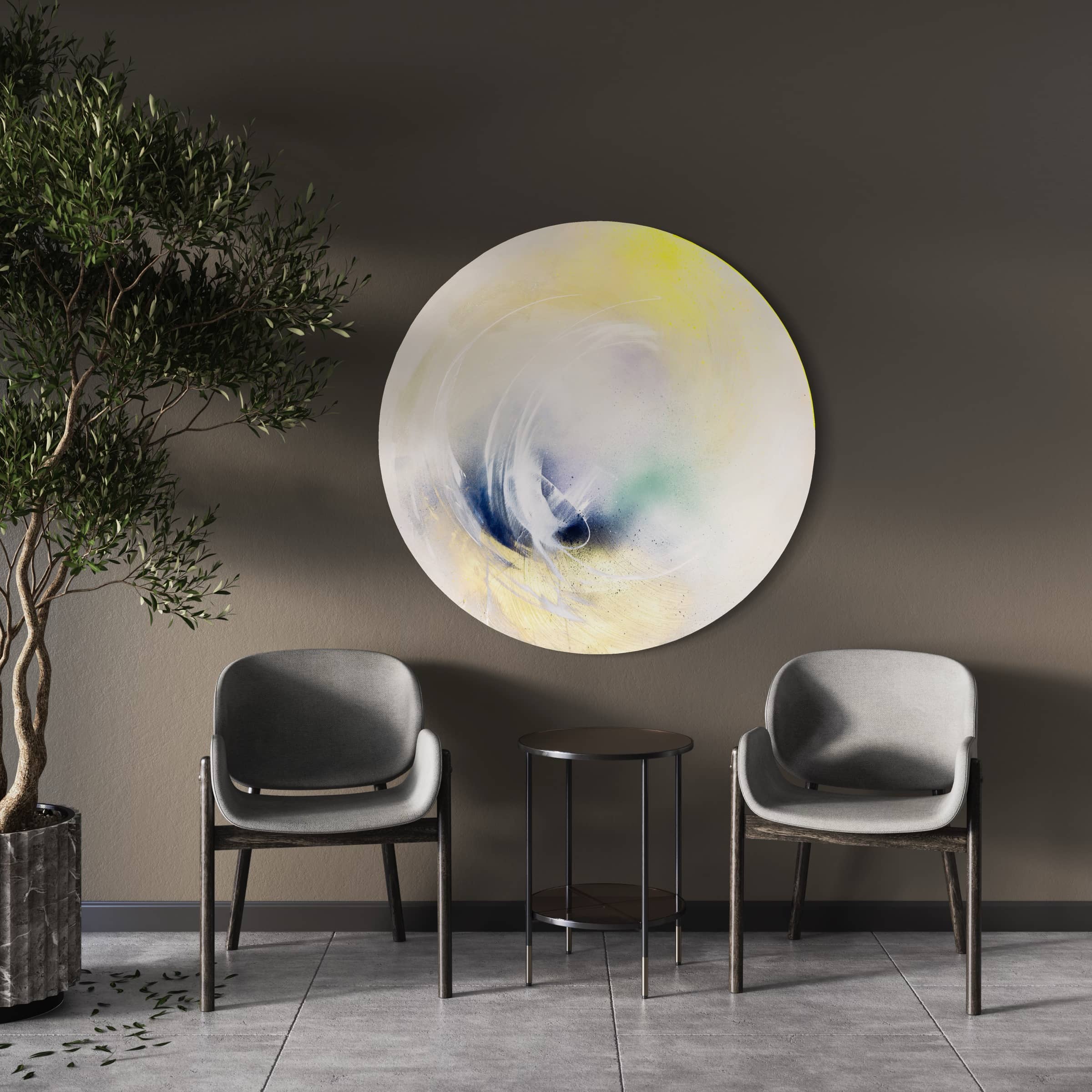 A minimalist room featuring two gray chairs and a round black table highlights "Let's Make This Moment Last," an original abstract painting by Kate Mayer, crafted with acrylic and spray paint, enhanced by a potted tree.