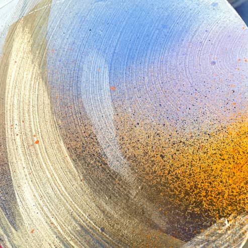 Title: "Be Joy" by Kate Mayer featuring swirling patterns of gold, blue, and orange in acrylic and spray paint. It showcases a textured gradient enriched with scattered specks.