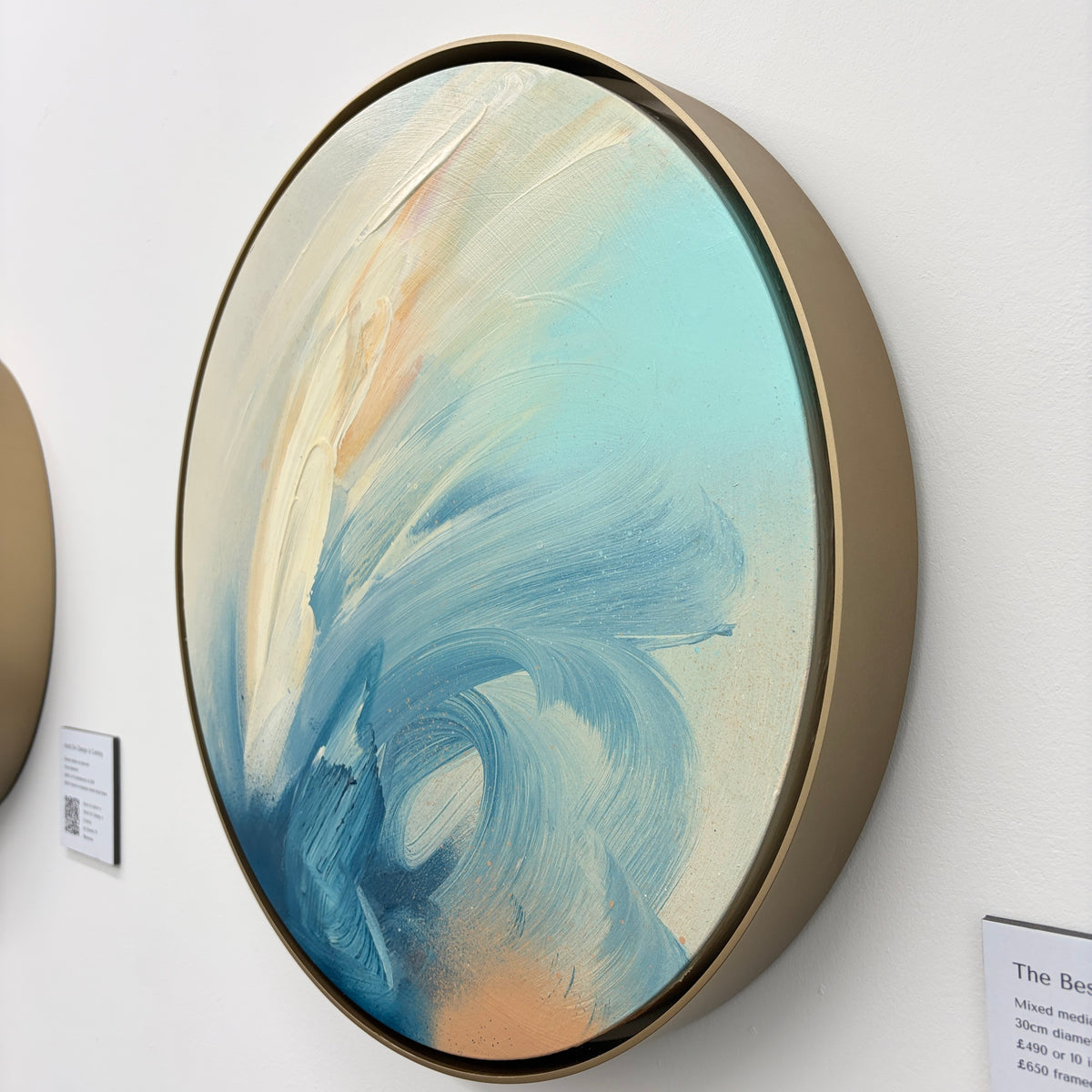 The Best Is Yet To Come“ by Kate Mayer: round blue and cream abstract acrylic on plywood in a tan frame.