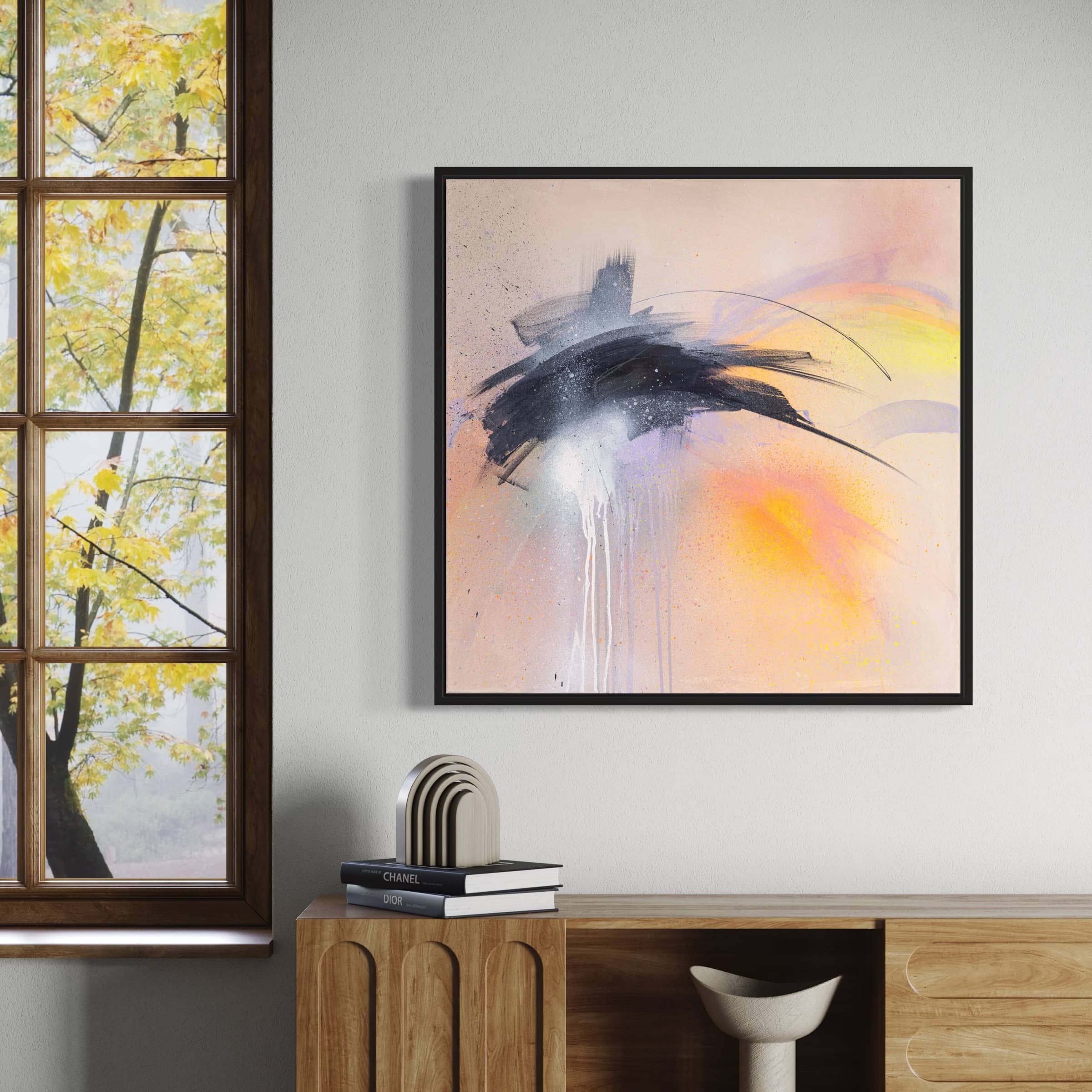 An abstract painting titled "That Moment On Black Sand Beach" by Kate Mayer, featuring black, white, and yellow splashes, hangs on a wall near a wooden cabinet and a window displaying autumn leaves. This original piece, crafted with acrylic and spray paint, includes a certificate of authenticity.