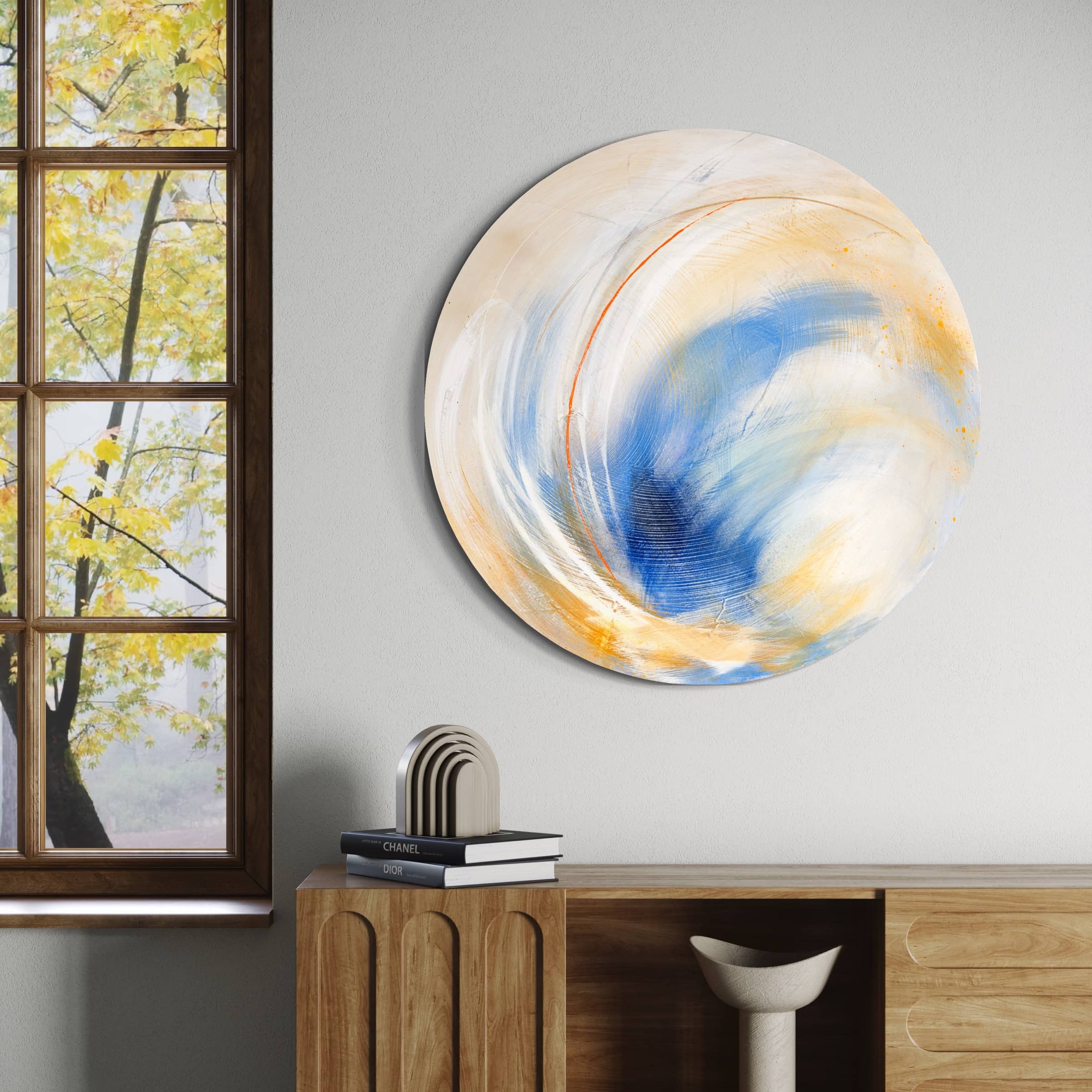 The "Go Your Own Way" original painting by Kate Mayer showcases vibrant blue and yellow swirls in a round abstract form. Crafted using acrylic and spray paint, it is ideally displayed on a wall next to a window that frames the view of autumn leaves, and above a finely crafted wooden cabinet.