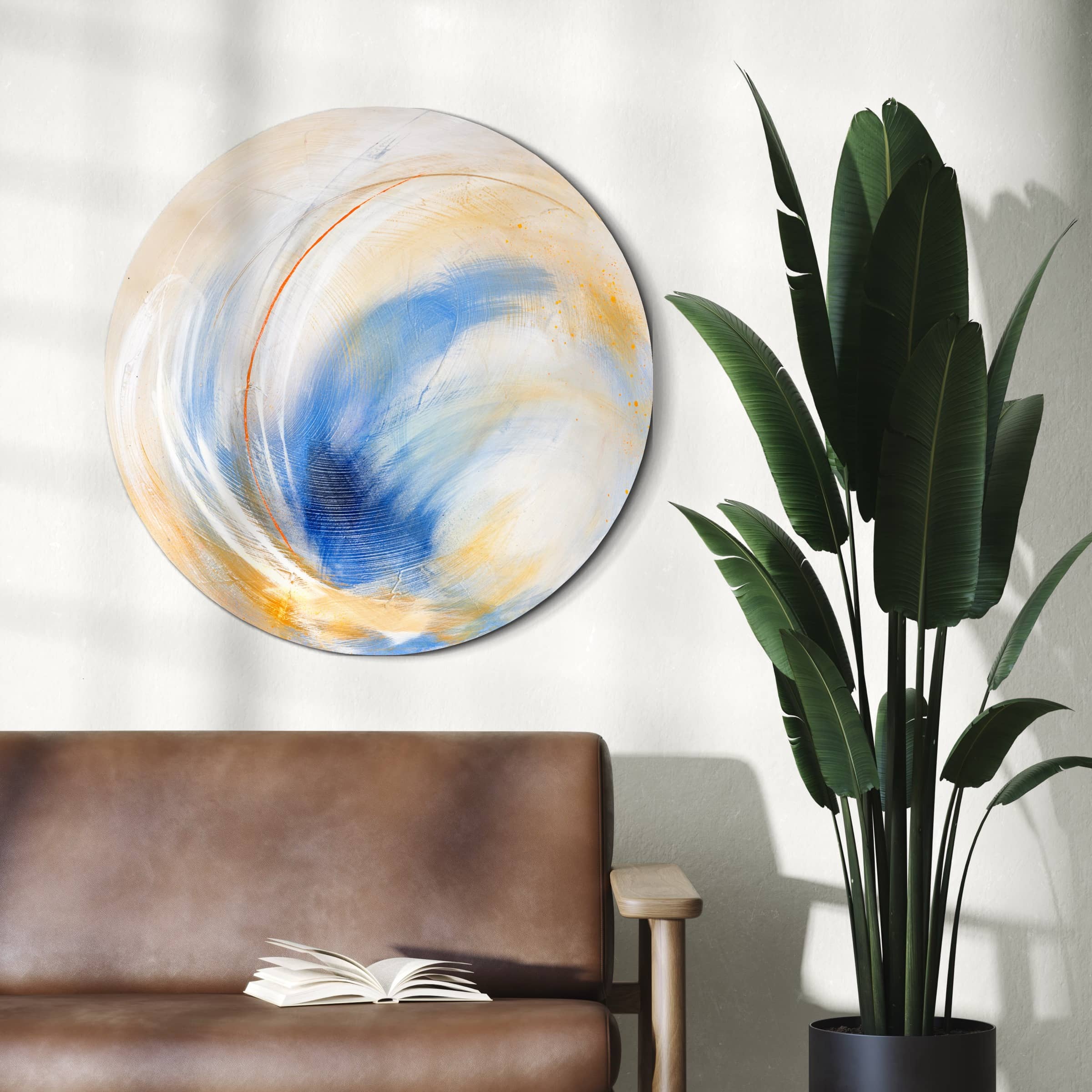 The "Go Your Own Way - Original Painting" by Kate Mayer, created with acrylic and spray paint, is displayed above a brown sofa. Its circular design in shades of blue, yellow, and white harmonizes with the adjacent green plant, while an open book lies enticingly on the sofa.