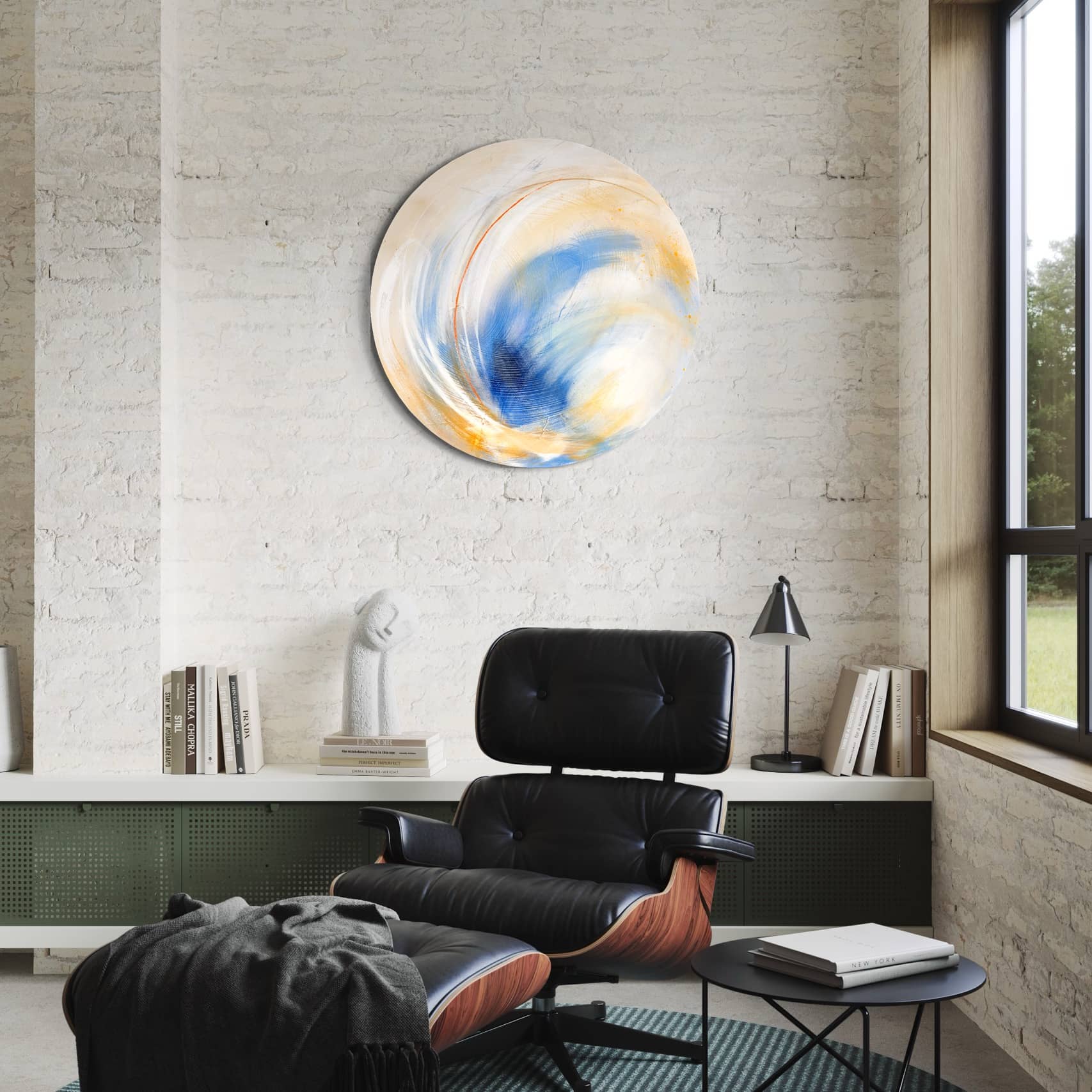 A modern lounge features a sleek black leather chair and a captivating piece of art, "Go Your Own Way" by Kate Mayer, mounted on the brick wall. This round abstract painting is crafted with acrylic and spray paint. Below the large window, an inviting shelf filled with books enhances the room's charm.