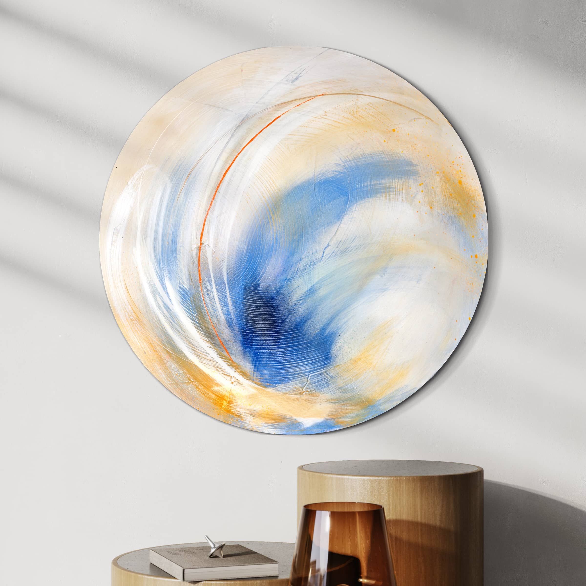 The round abstract artwork, titled "Go Your Own Way - Original Painting" by Kate Mayer, showcases swirling blue, white, and orange patterns created with acrylic and spray paint. It is displayed on a wall near a stack of books and a glass.