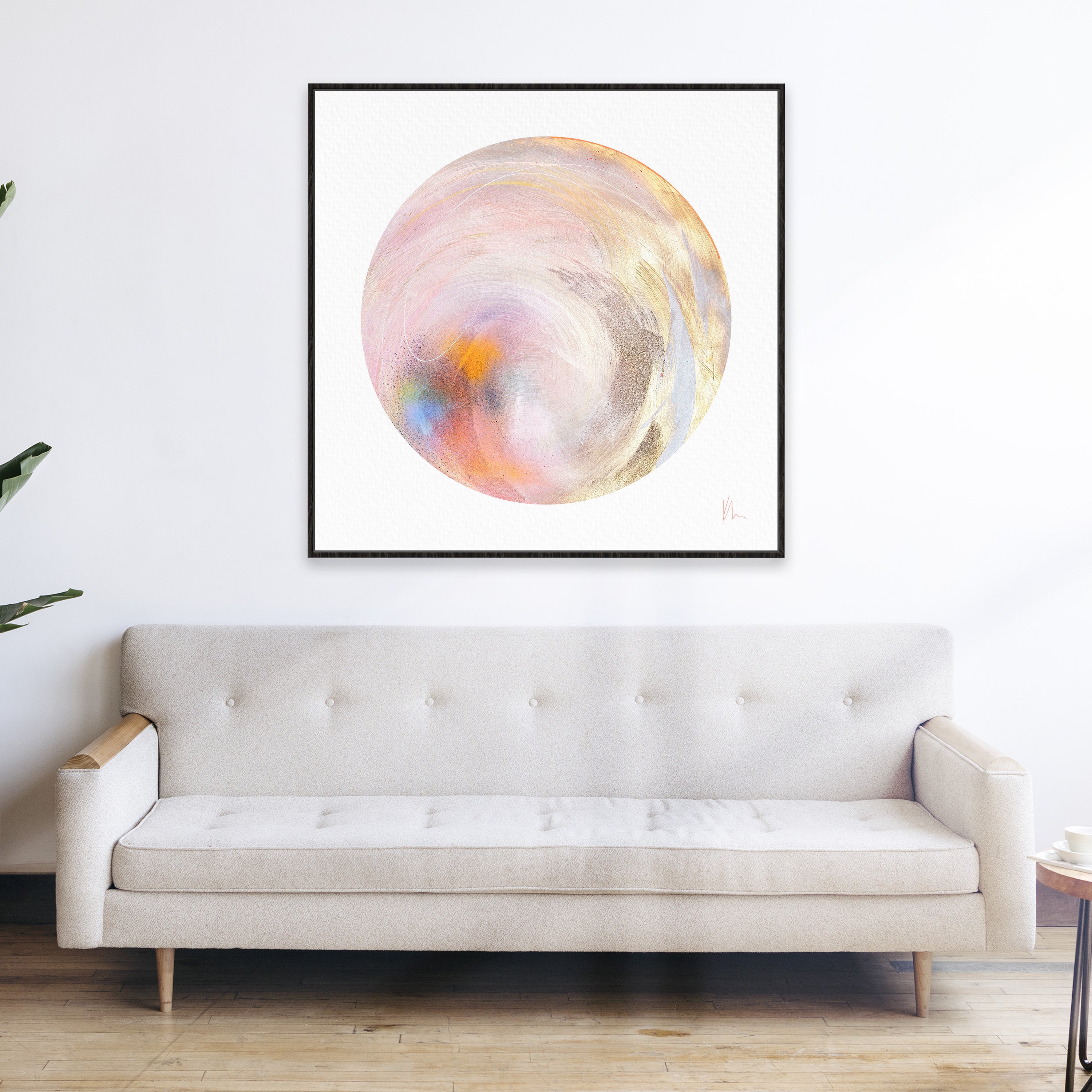 The "Believe In Me" artwork by Kate Mayer, featuring abstract circular designs with pastel hues adorned with metallic gold spray paint, is displayed above a beige sofa in a minimalist room.