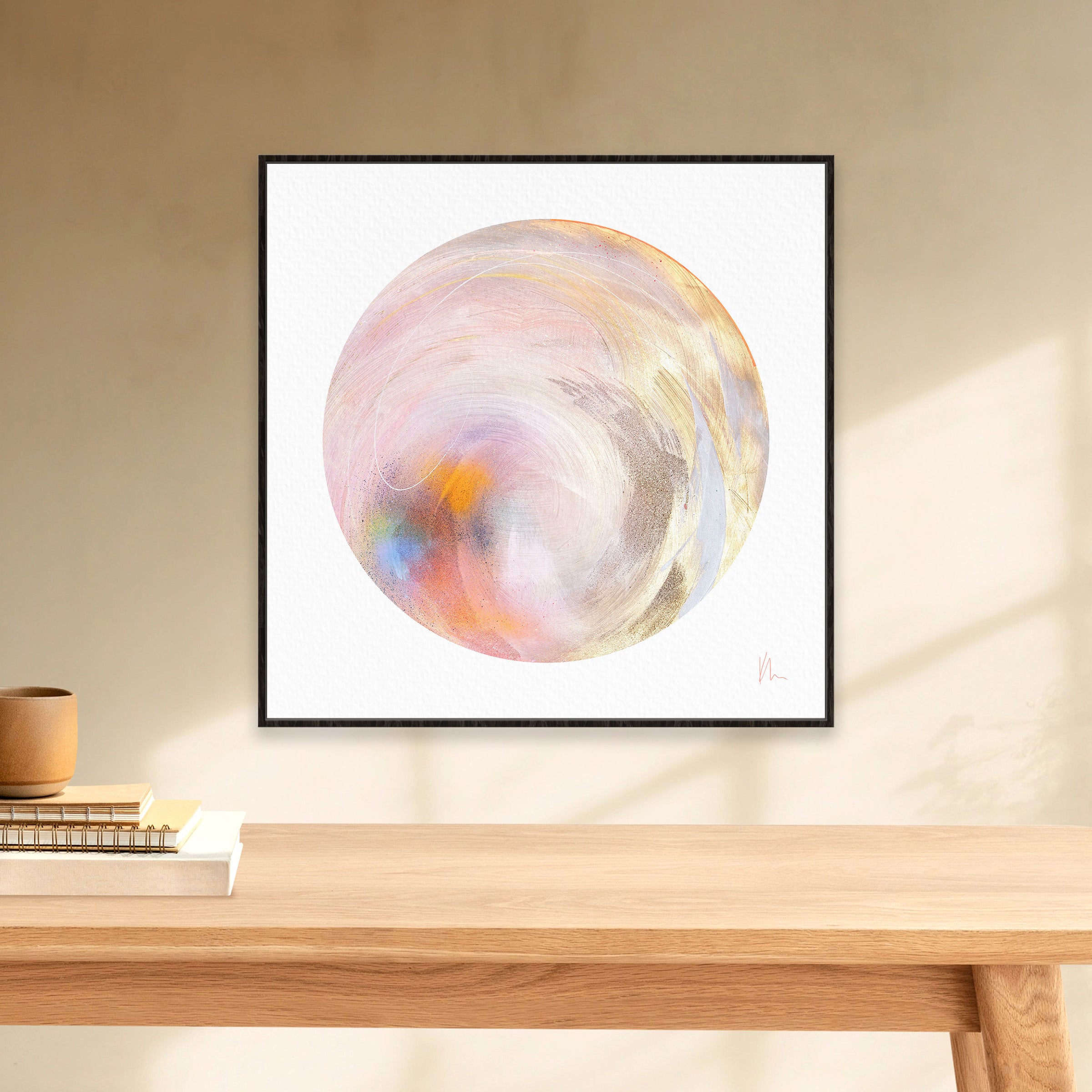 The "Believe In Me" artwork by Kate Mayer showcases abstract circular pastel swirls with a touch of metallic gold spray paint, elegantly displayed above a wooden table adorned with books and a ceramic pot.