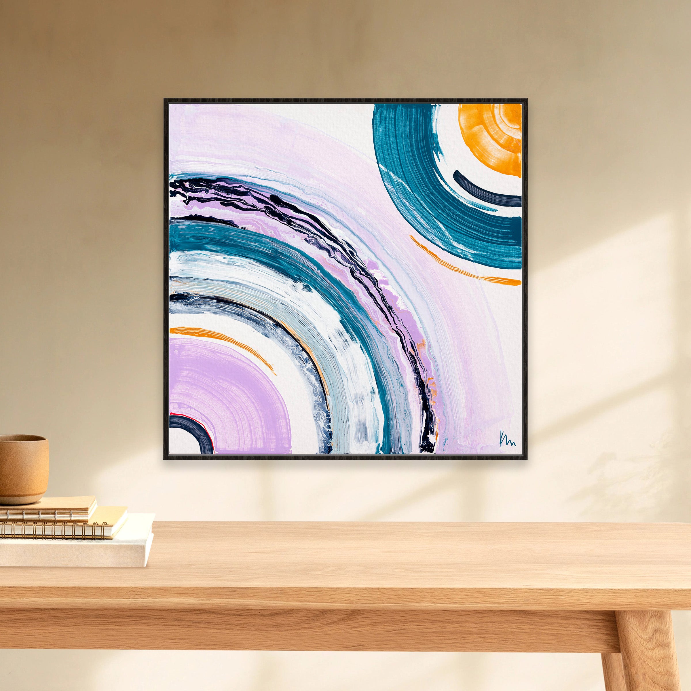 Framed inspiring artwork titled "Between Us" by Kate Mayer, featuring colorful circular patterns, adorns a light wall above a wooden table. Nearby, books and a small vase rest beneath the abstract print, evoking a sense of human connection.