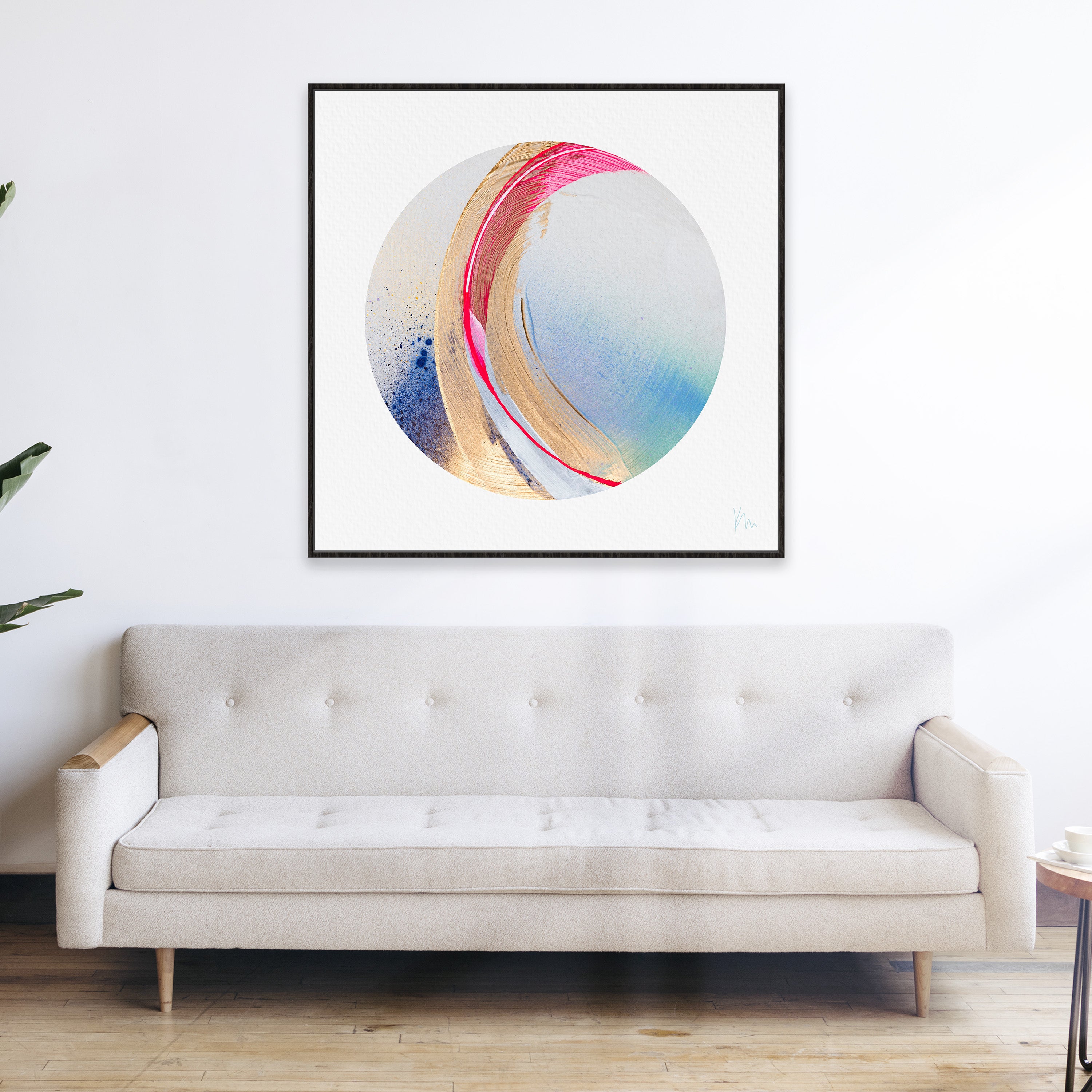 A piece titled "Be Radiant" by Kate Mayer, featuring an abstract circular artwork with vibrant colors, is elegantly displayed above a beige sofa in a minimalist room with wooden flooring.