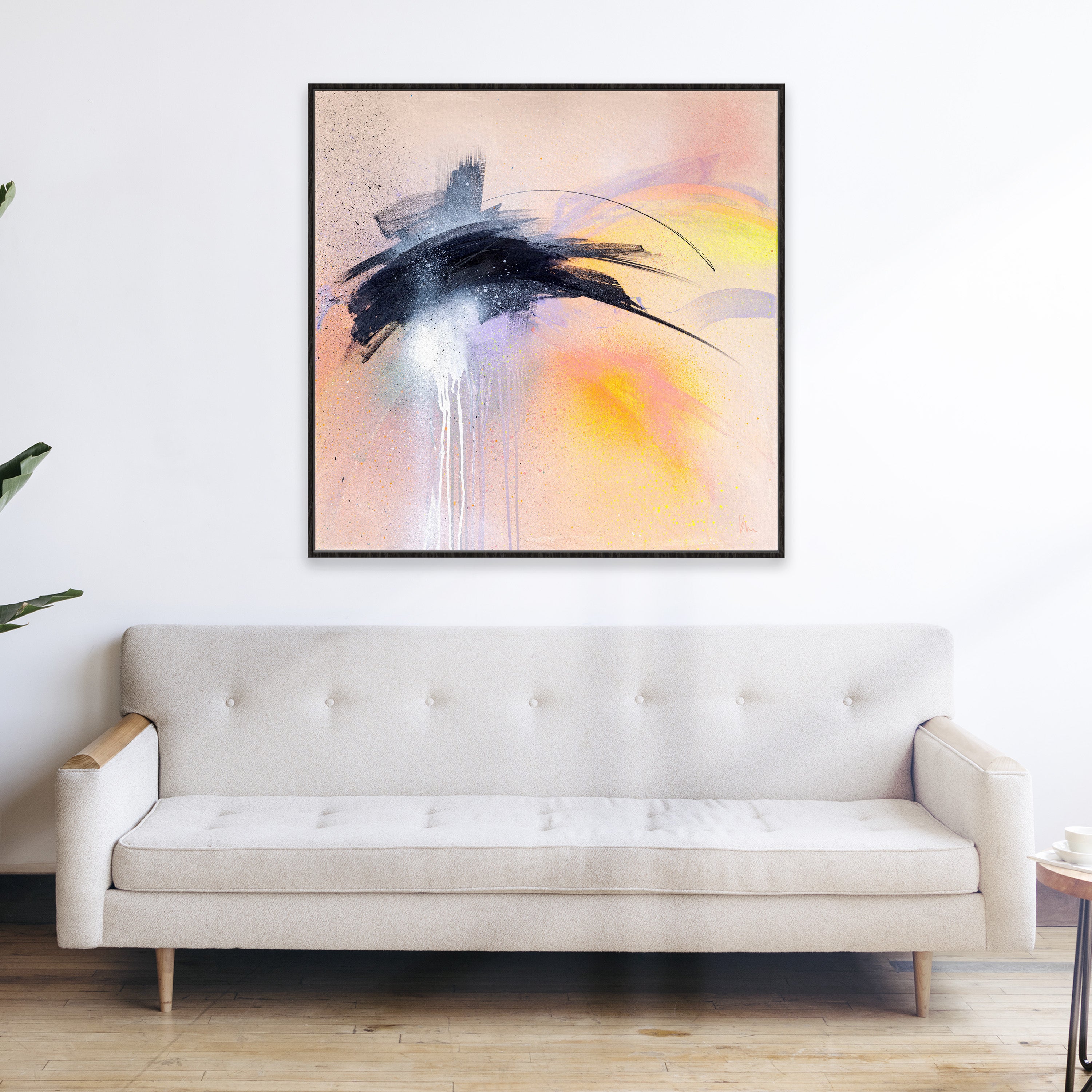 A beige sofa sits against a white wall, adorned with "Black Sand Beach," a vibrant abstract painting by Kate Mayer that captures the essence of a sunset over Iceland, featuring expressive black, white, and yellow strokes hanging above.