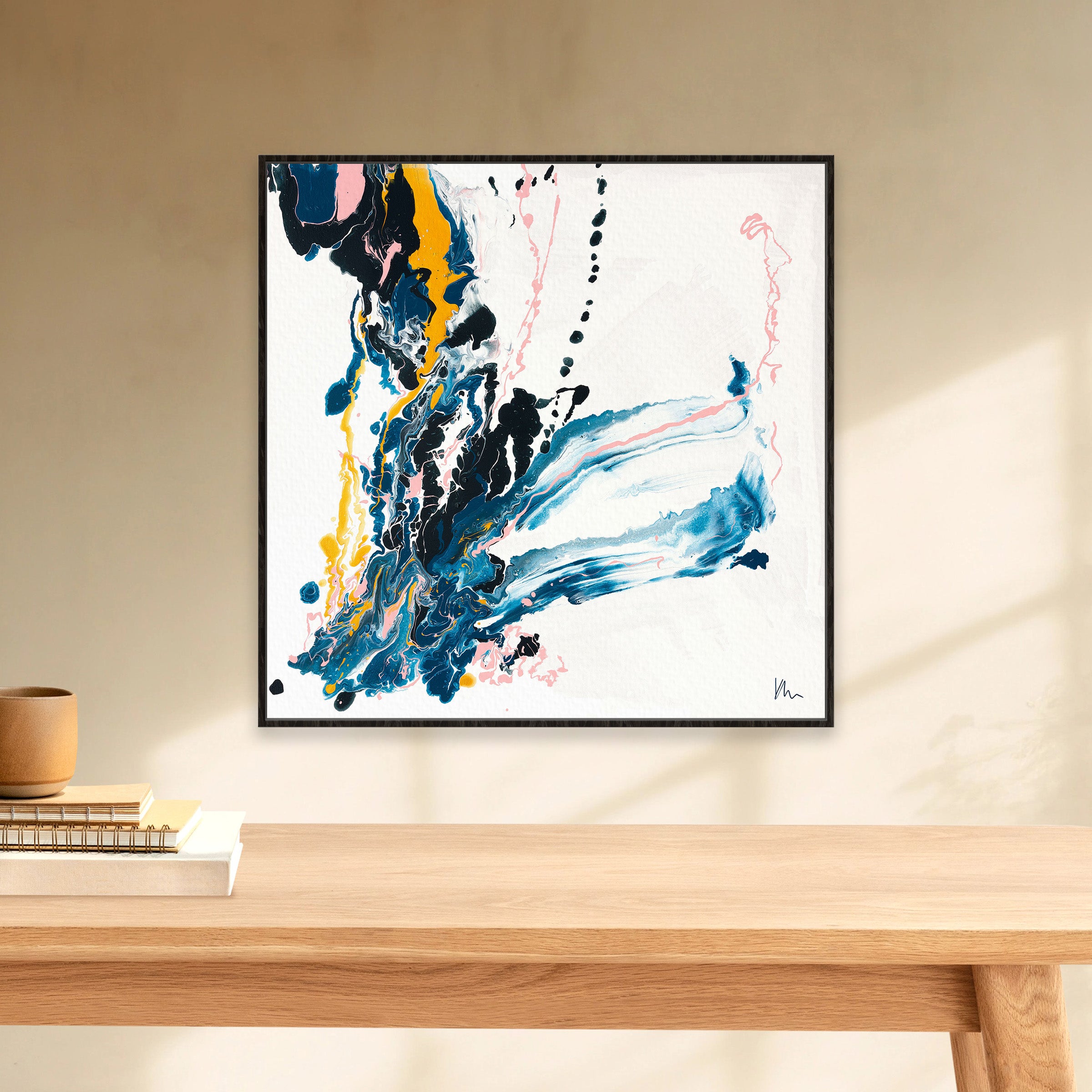 Kate Mayer's "Emerging," an abstract painting featuring vibrant blue, yellow, and pink brushstrokes that symbolize new beginnings, is mounted on a beige wall above a wooden table adorned with books.