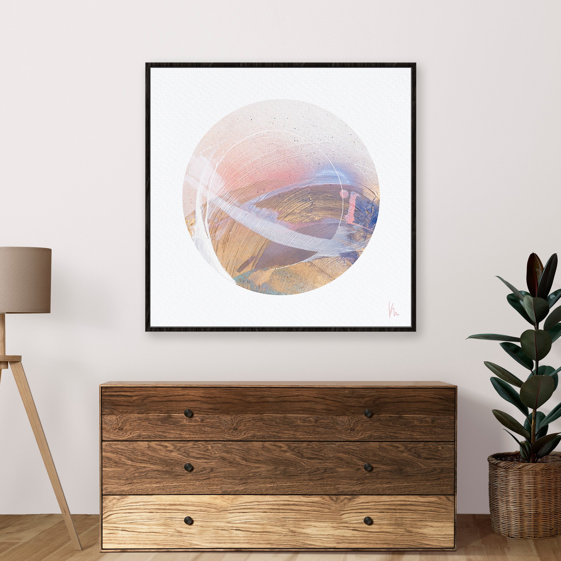 A modern abstract circular painting titled "Just Be" by Kate Mayer, evoking inner harmony, is displayed above a wooden dresser with three drawers, accompanied by a floor lamp and a potted plant.