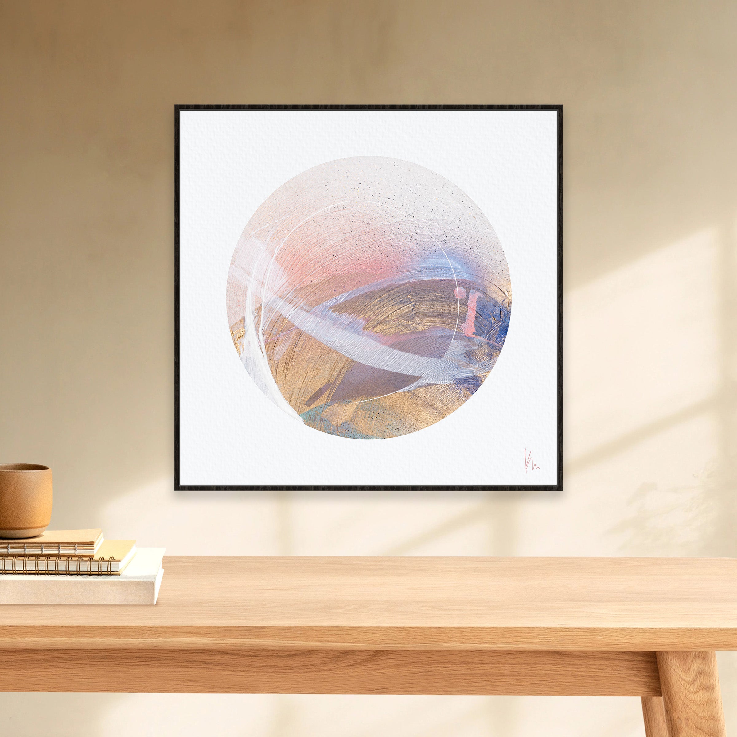 The "Just Be" framed abstract circular artwork by Kate Mayer, with its pastel and earthy tones, evokes inner harmony when placed on a beige wall above a wooden table adorned with books and a small cup.