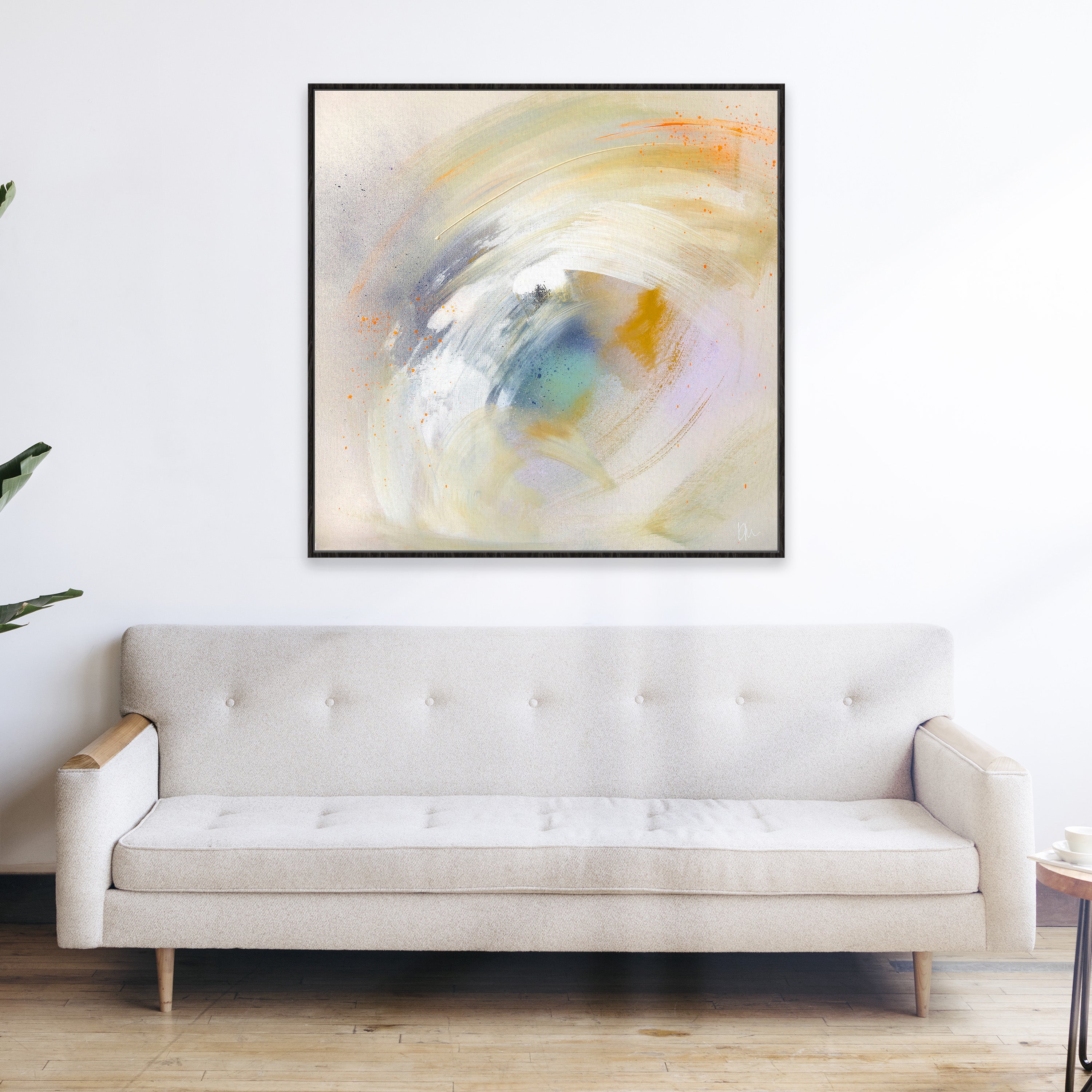 Make A Wish," an abstract painting by Kate Mayer featuring swirls of white, yellow, and blue, adorns the wall above a beige sofa on a wooden floor. This charming addition captures an inviting atmosphere perfect for any living space.