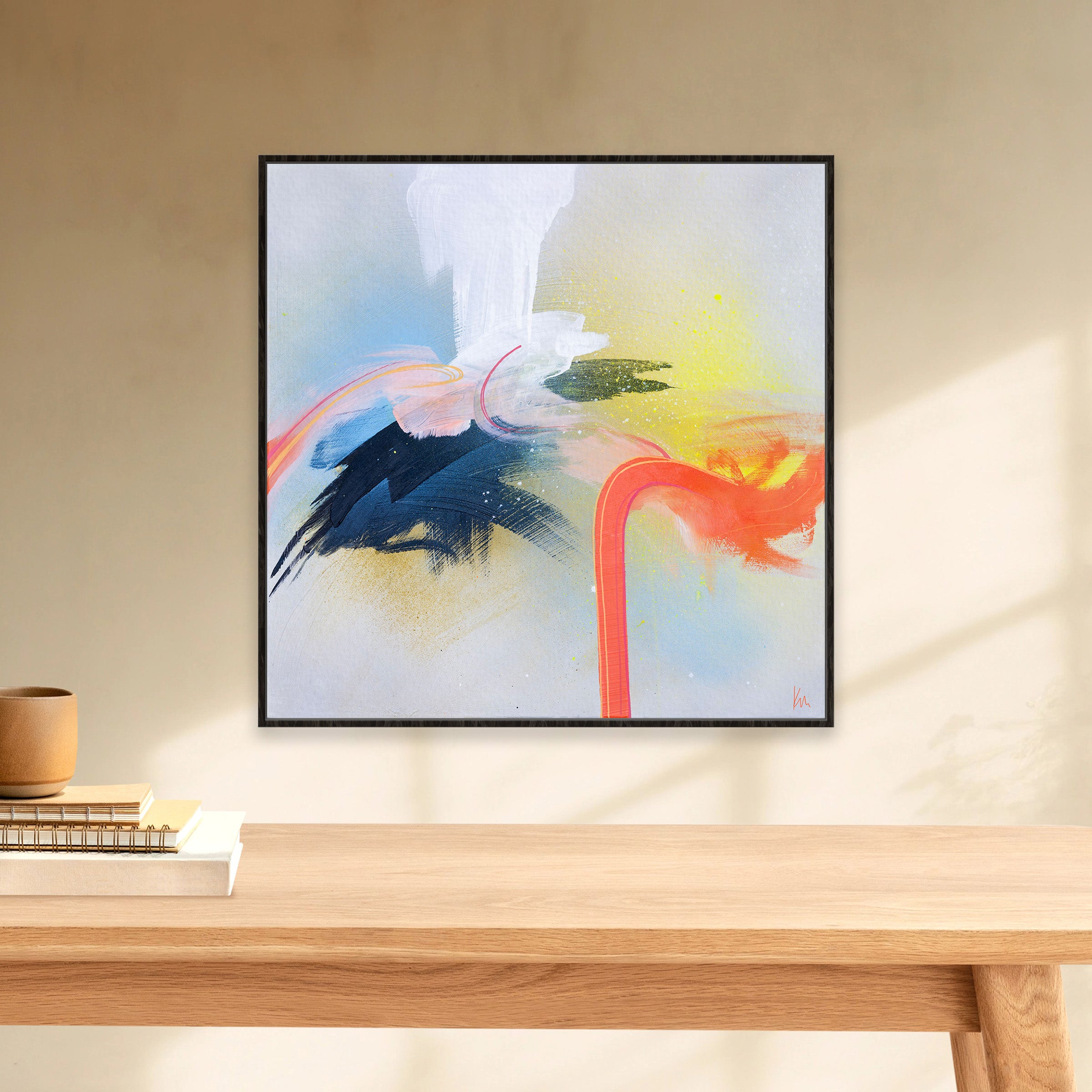 The "Miami Flamingo" painting by Kate Mayer, featuring vibrant abstract brushstrokes of blue, orange, and yellow, enhances the wall above a wooden table decorated with books and a cup, creating an inviting artistic atmosphere.