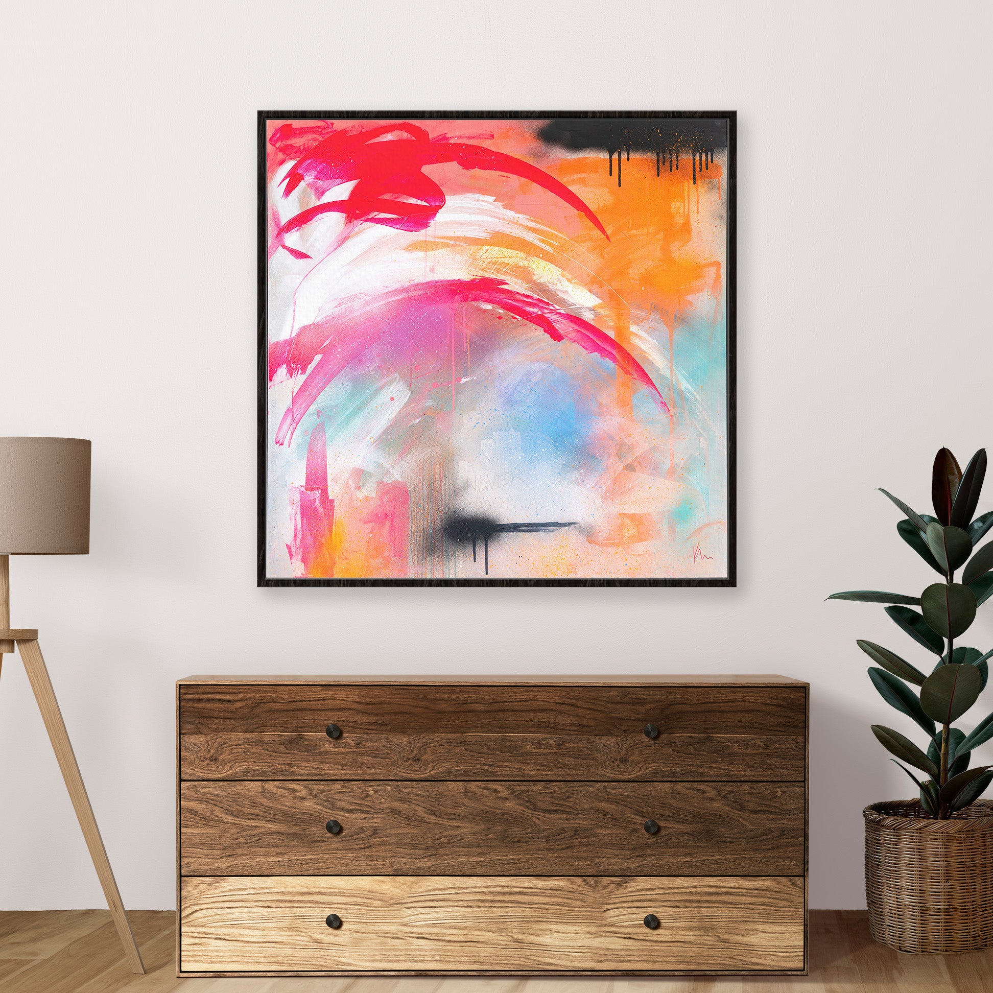 Kate Mayer's 'Never Too Much' abstract painting, featuring vibrant pink, orange, and blue strokes, embodies boldness as it hangs above a wooden dresser. Flanked by a lamp and plant, it serves as a visual reminder that when it comes to art, there can never be too much expression.
