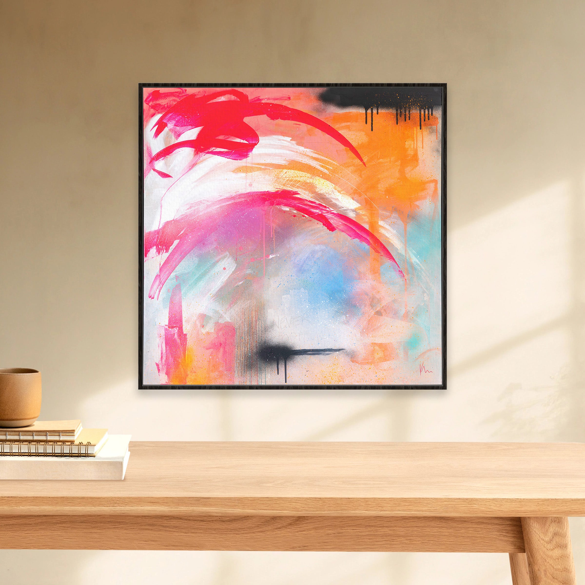 A vibrant painting titled "Never Too Much" by Kate Mayer showcases dynamic brushstrokes in red, orange, pink, and blue. It hangs on a beige wall above a wooden table, embodying the notion that in art and creativity, there's an endless embrace of color and expression.