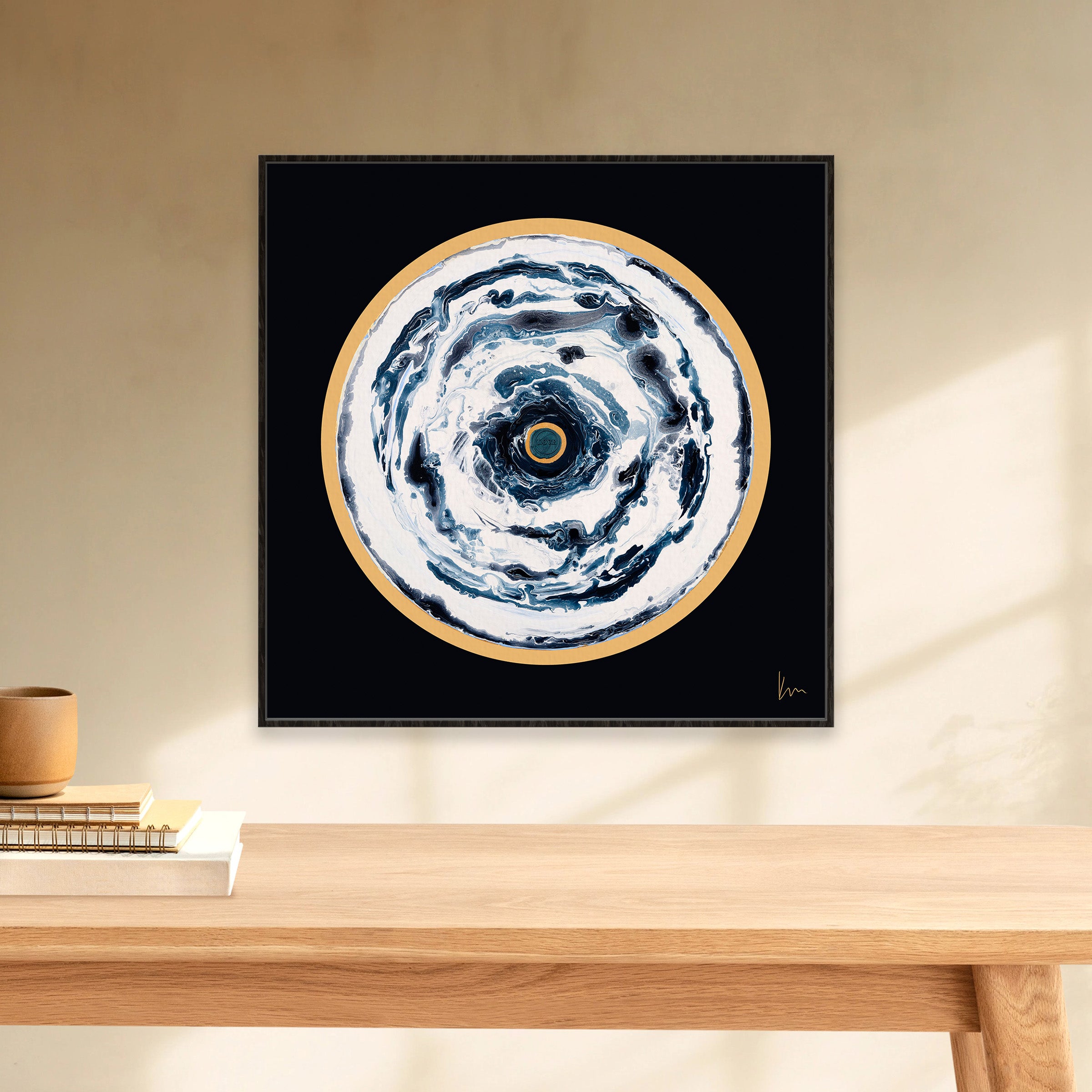 The "Oceans Of Love" painting by Kate Mayer, featuring abstract circular designs in black, white, and gold tones, graces the wall above a wooden table adorned with books and a delicate vase.