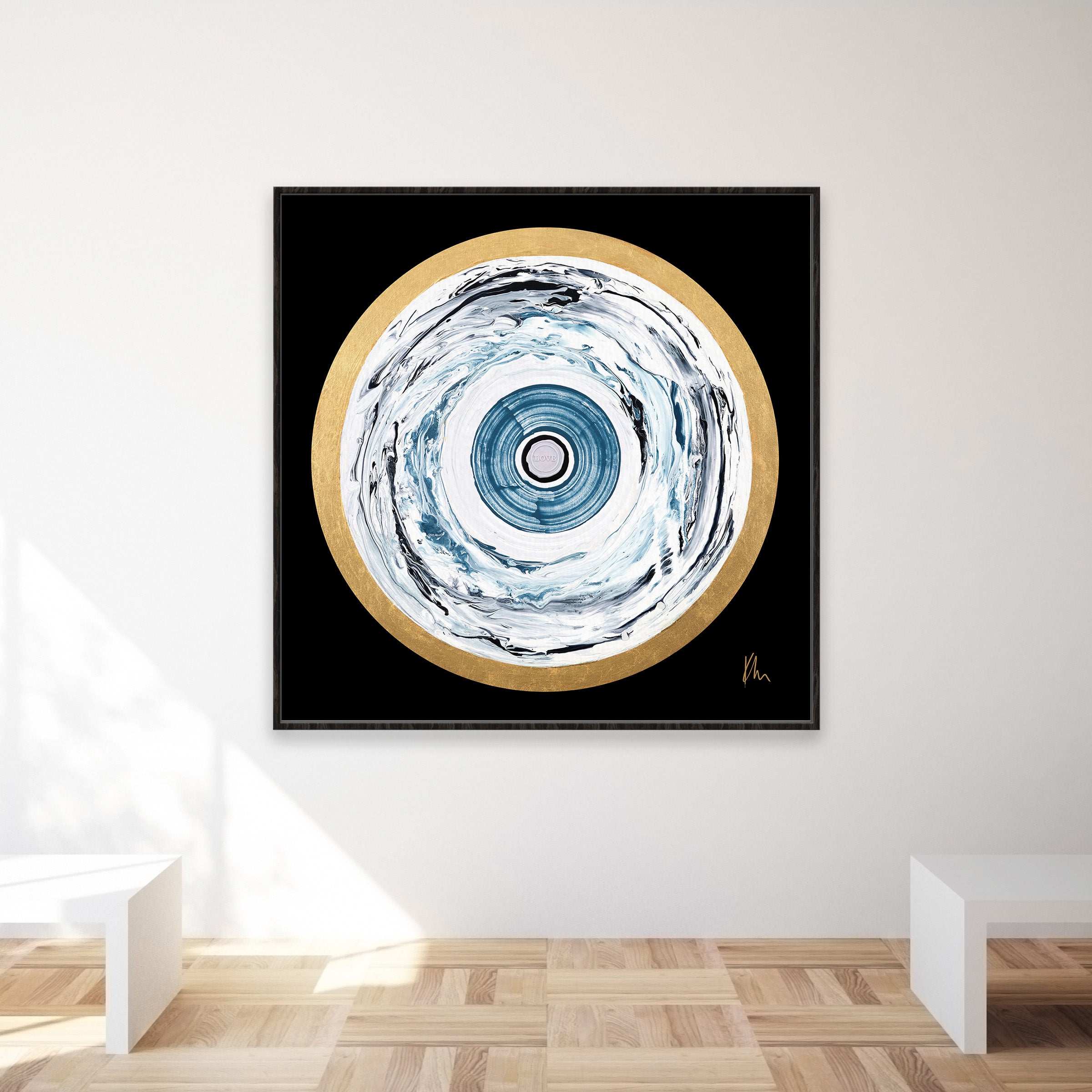 The "One Love" framed abstract painting by Kate Mayer, featuring concentric circles in blue, white, and gold, hangs elegantly on a minimalist white wall above a wooden floor.