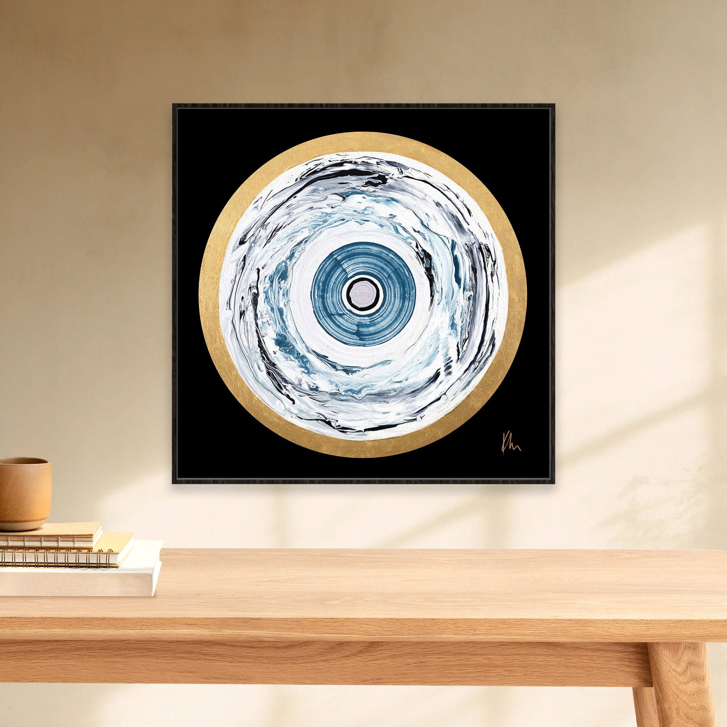 The "One Love" abstract painting by Kate Mayer, featuring concentric circles in blue, white, and gold on a black background, hangs elegantly above a wooden table. This piece exudes the essence of love through its harmonious blend of vibrant colors.