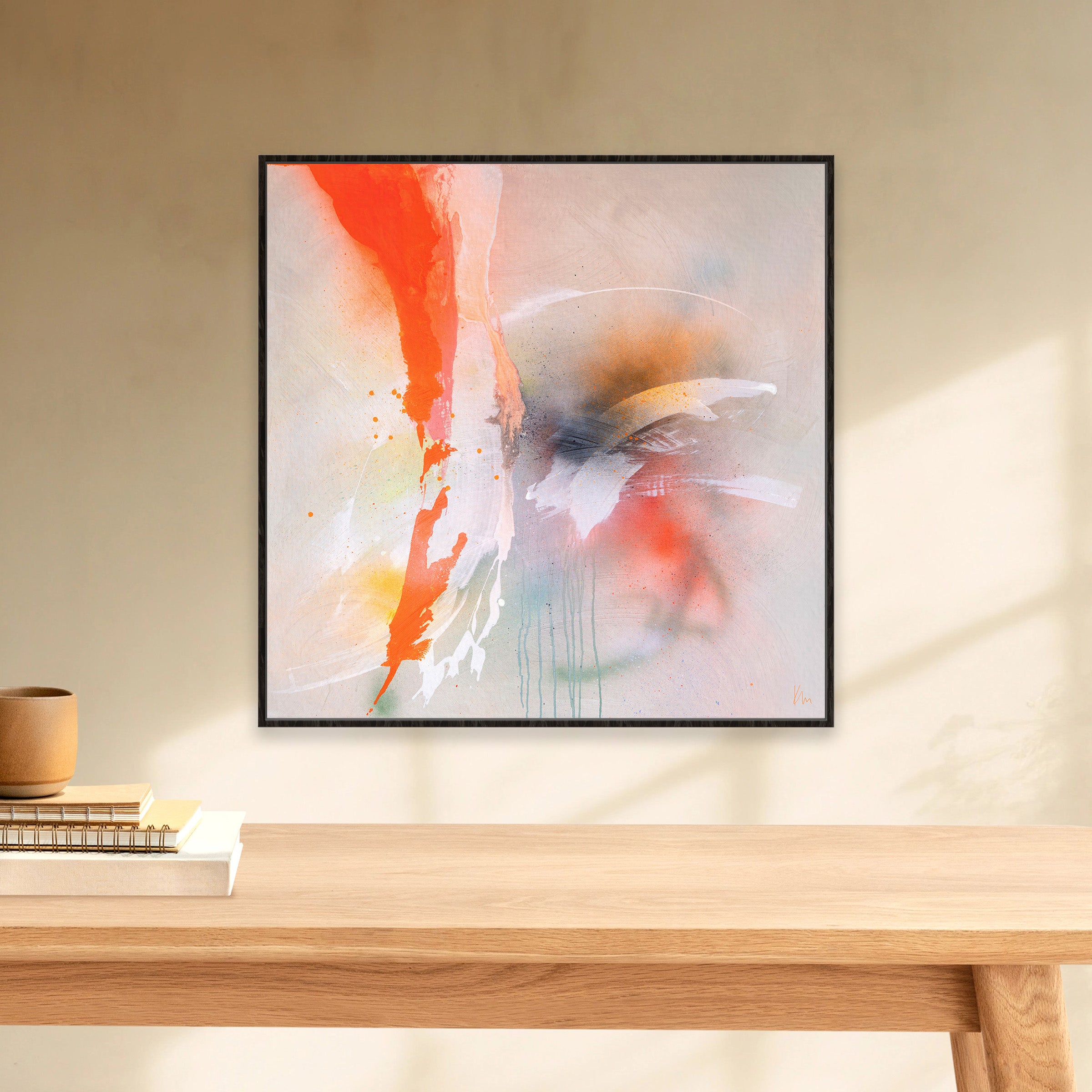 Kate Mayer's 'Rainbow Falls,' a modern abstract painting featuring red, orange, and white brushstrokes, hangs on a beige wall above a wooden table decorated with books and a ceramic cup.