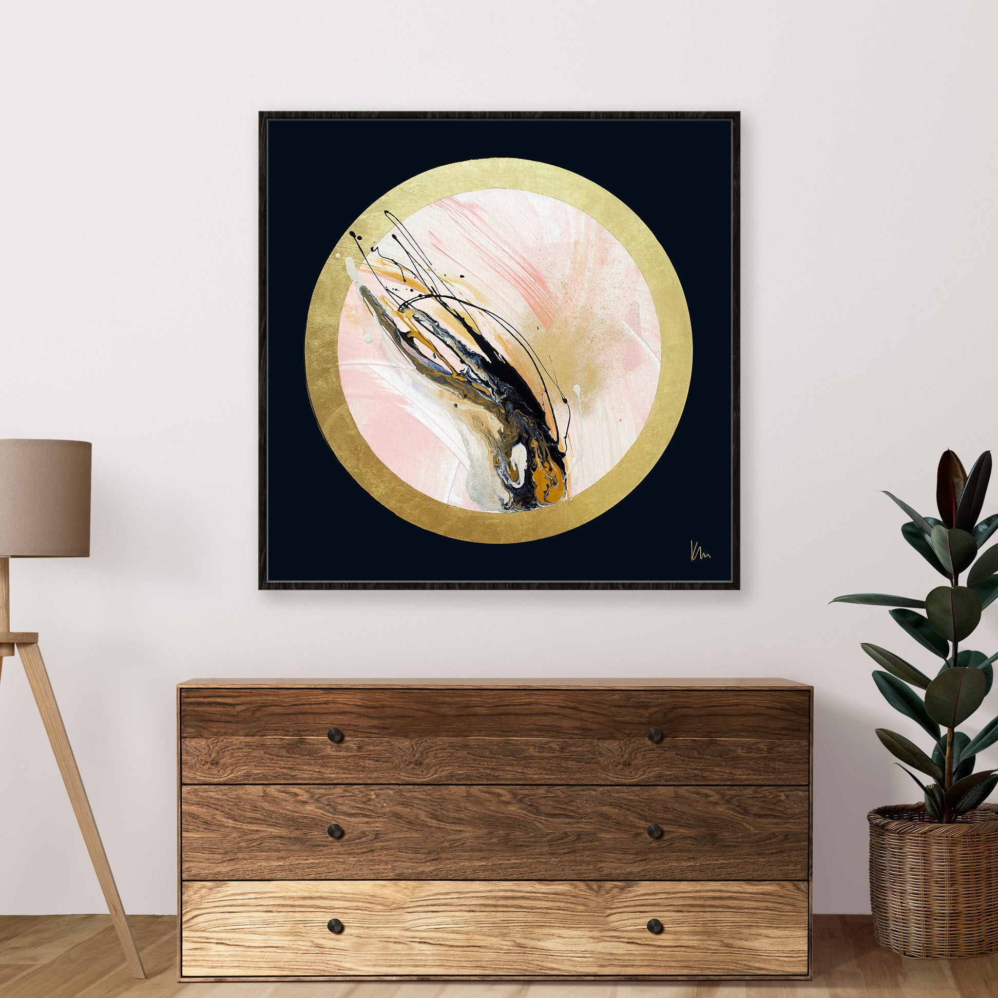 The "Rise Up" abstract painting by Kate Mayer, featuring pink, black, and gold hues in a circle frame, hangs above a wooden chest. It's complemented by a potted plant and lamp nearby, capturing the elegant scene perfectly.