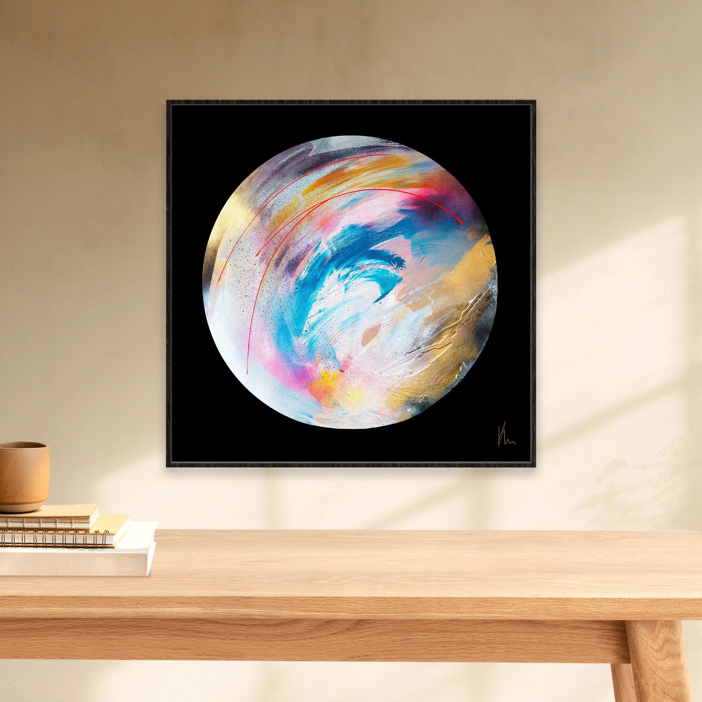 The "Speak Your Truth" abstract painting by Kate Mayer, featuring a colorful swirling circle, is beautifully showcased in a black frame. It hangs elegantly on a beige wall above a light wooden surface adorned with books and decor. This description captures the vivid essence of the artwork perfectly.